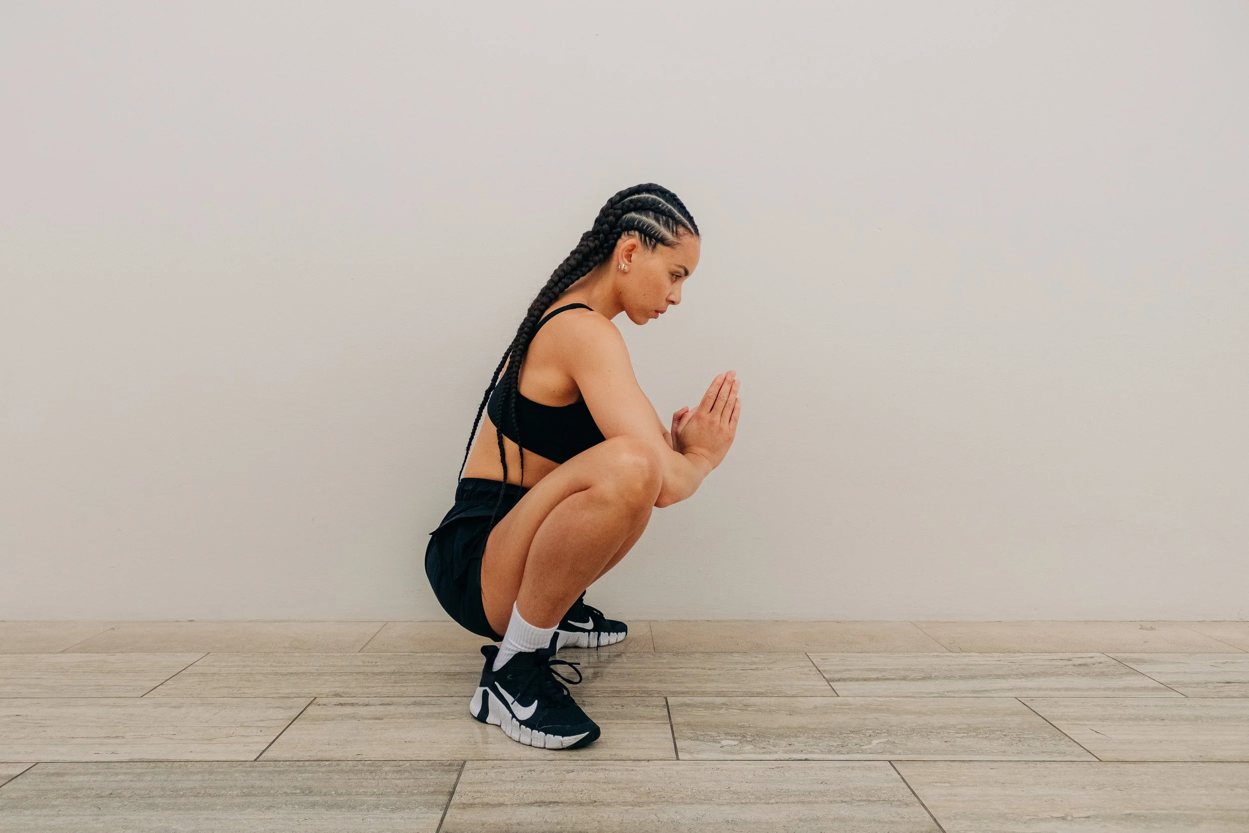 A woman with braided hair in a black sports bra and shorts is squatting on the floor with her hands pressed together in a prayer position, and her head bowed forward.