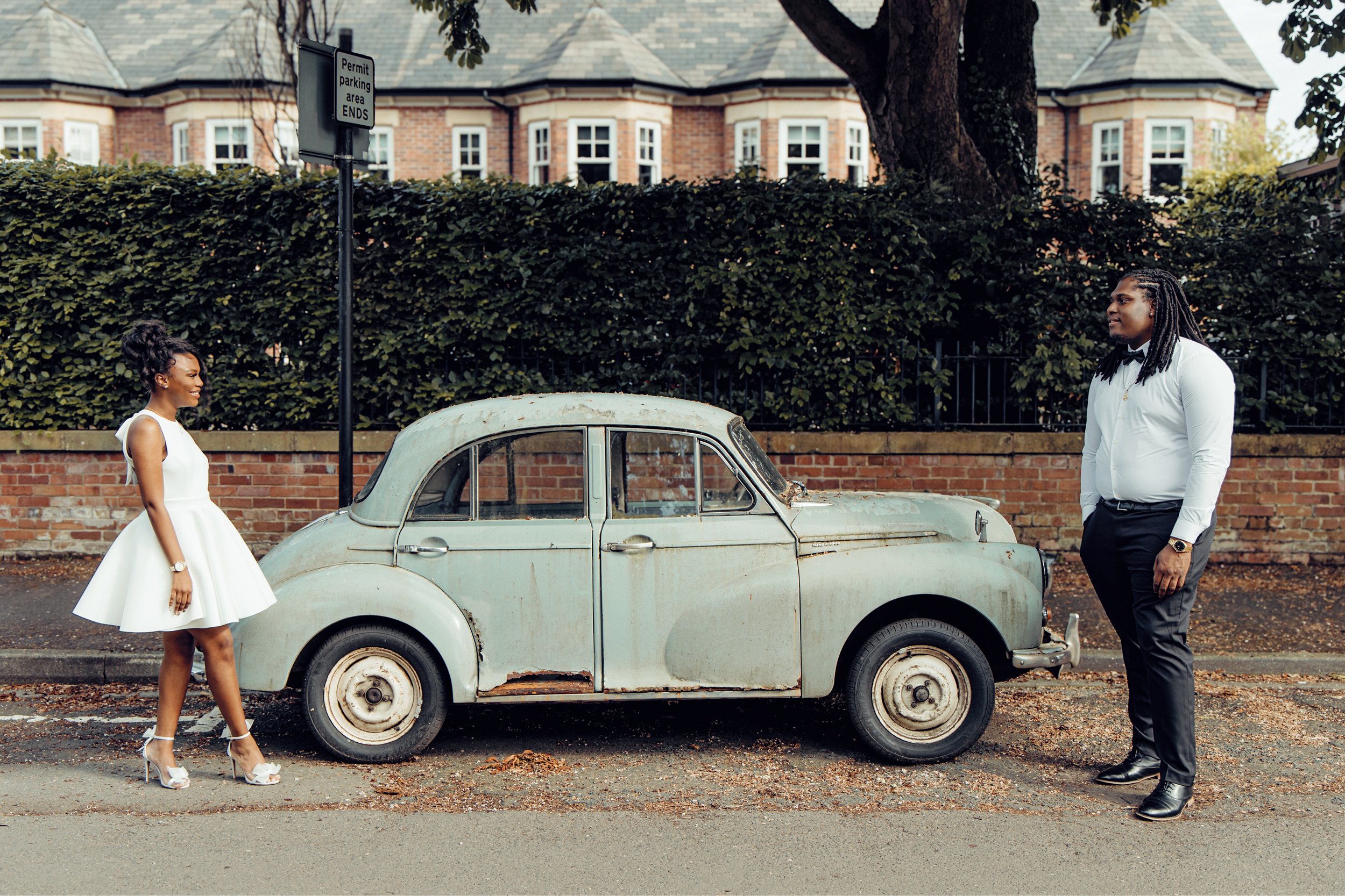 A man and woman dressed semi-formally are standing on opposite sides of a vintage, small, weathered car parked on the street. The woman is wearing a white dress and high heels, and the man is in a white shirt and dark trousers. They are facing each o