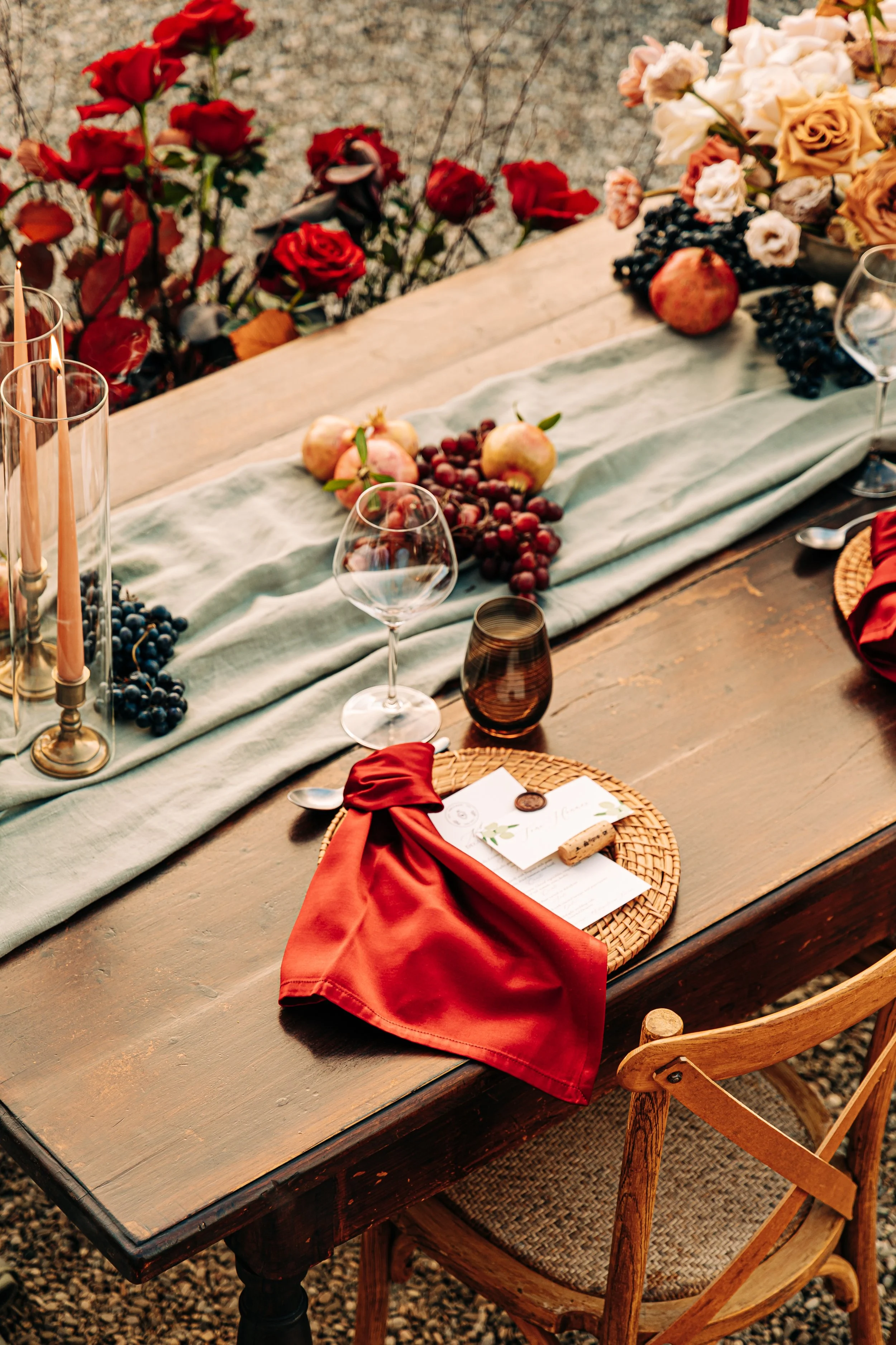 A rustic wooden table set outdoors with a light green table runner, surrounded by wooden chairs. The table is decorated with red apples, pomegranate, black grapes, white and pink roses, candles, wine glasses, and a red cloth napkin.