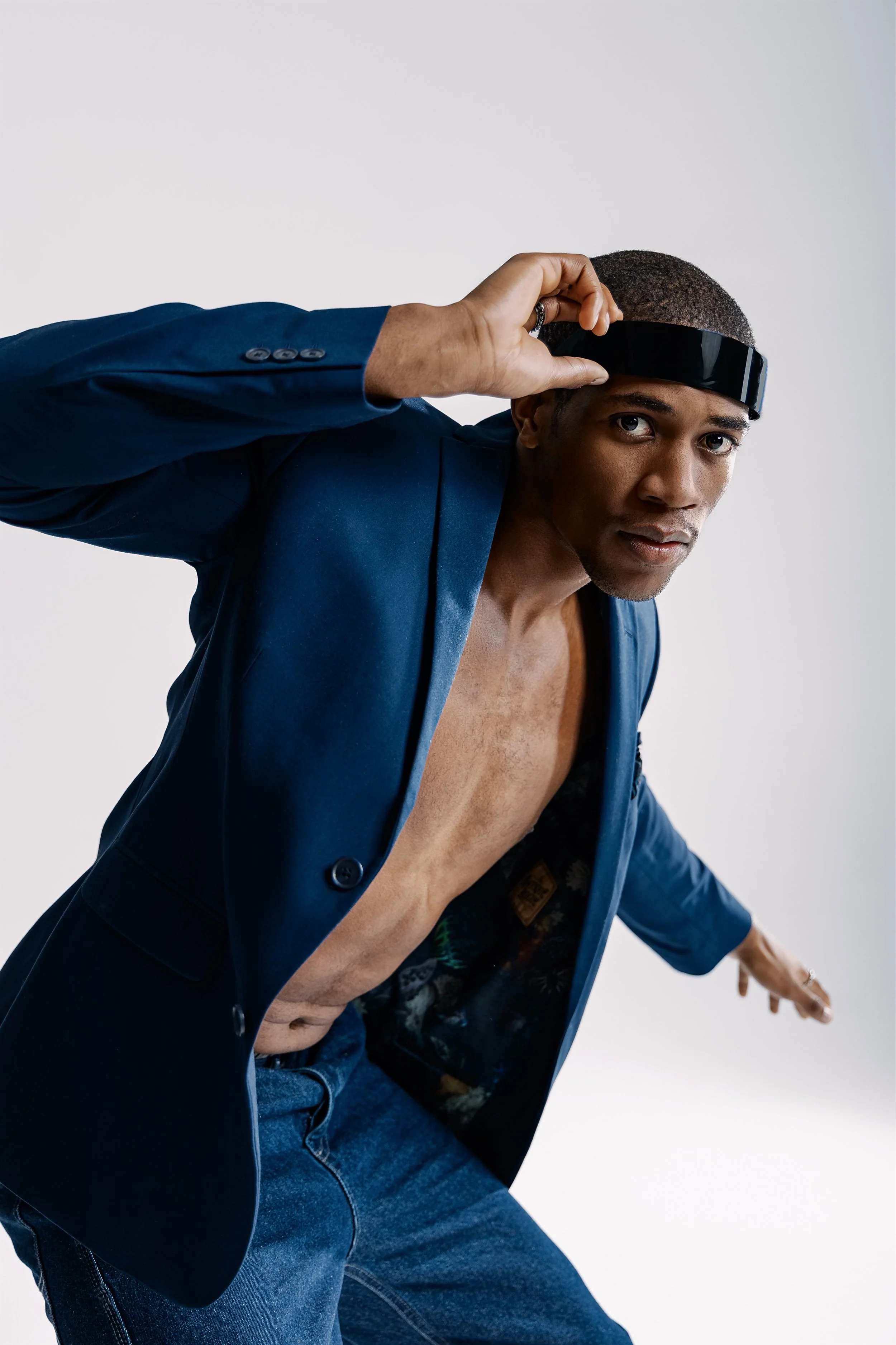 A man wearing a blue blazer, jeans, and a black visor, posing against a plain background.