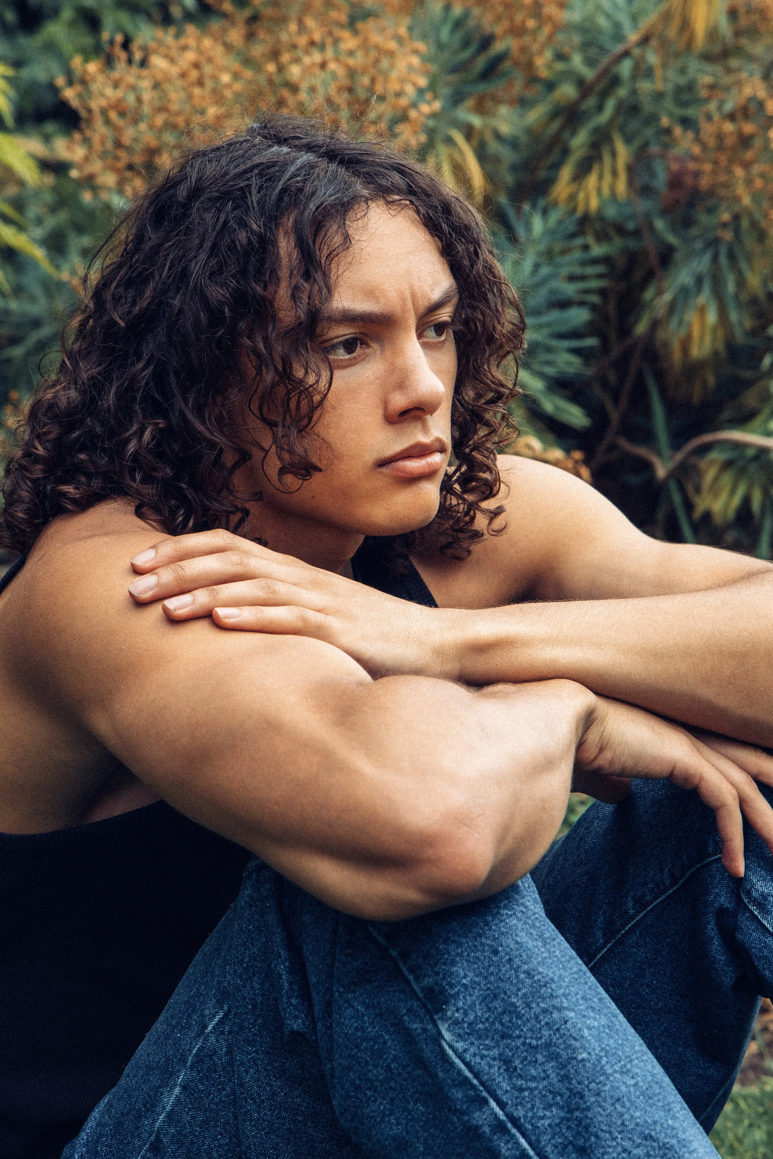 A woman with curly dark hair sitting outdoors, resting her arms on her knees, surrounded by green foliage.
