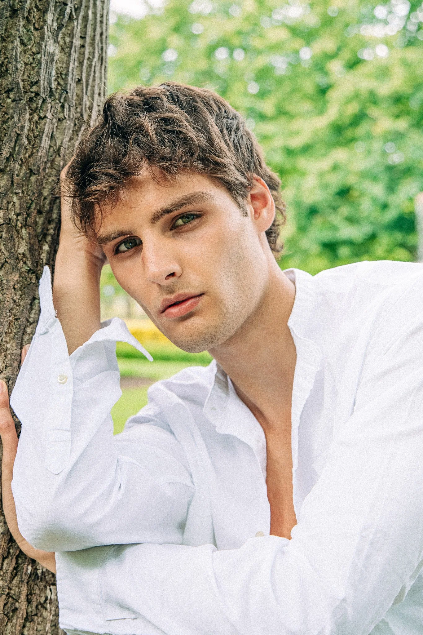 Young man with wavy brown hair and green eyes in a white shirt leaning against a tree outdoors with a blurred green background.