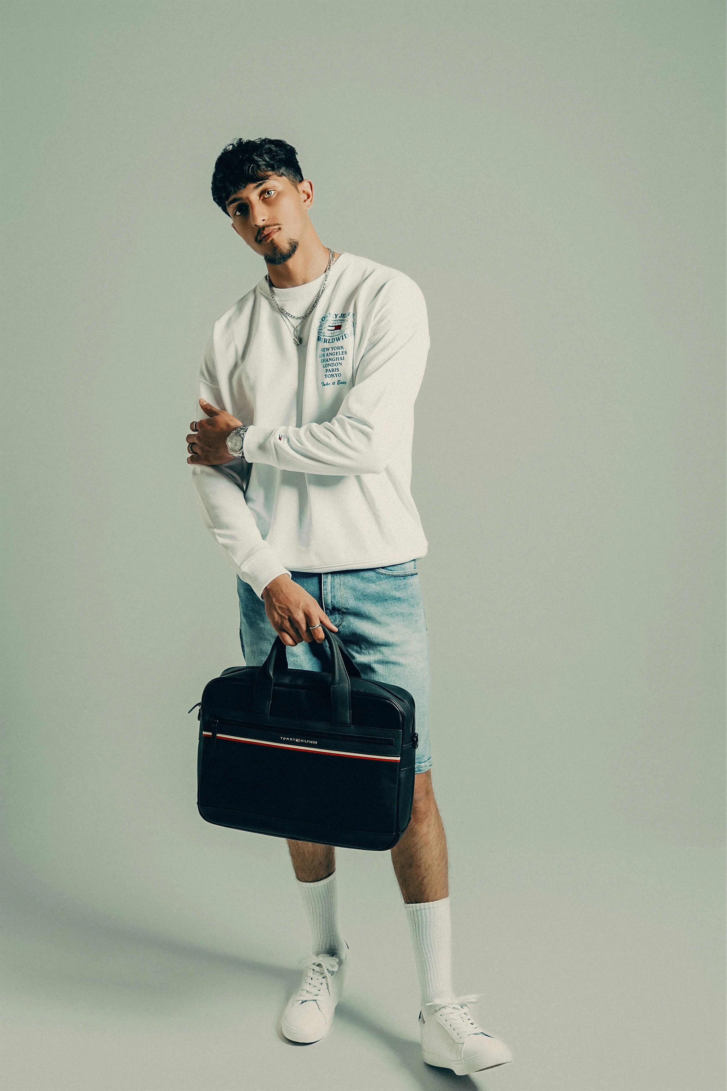 A young man with dark hair and a goatee stands against a plain backdrop, holding a black Tommy Hilfiger briefcase in his right hand. He wears a long-sleeve white shirt with blue text and graphics, light denim shorts, white sneakers, and white socks. 