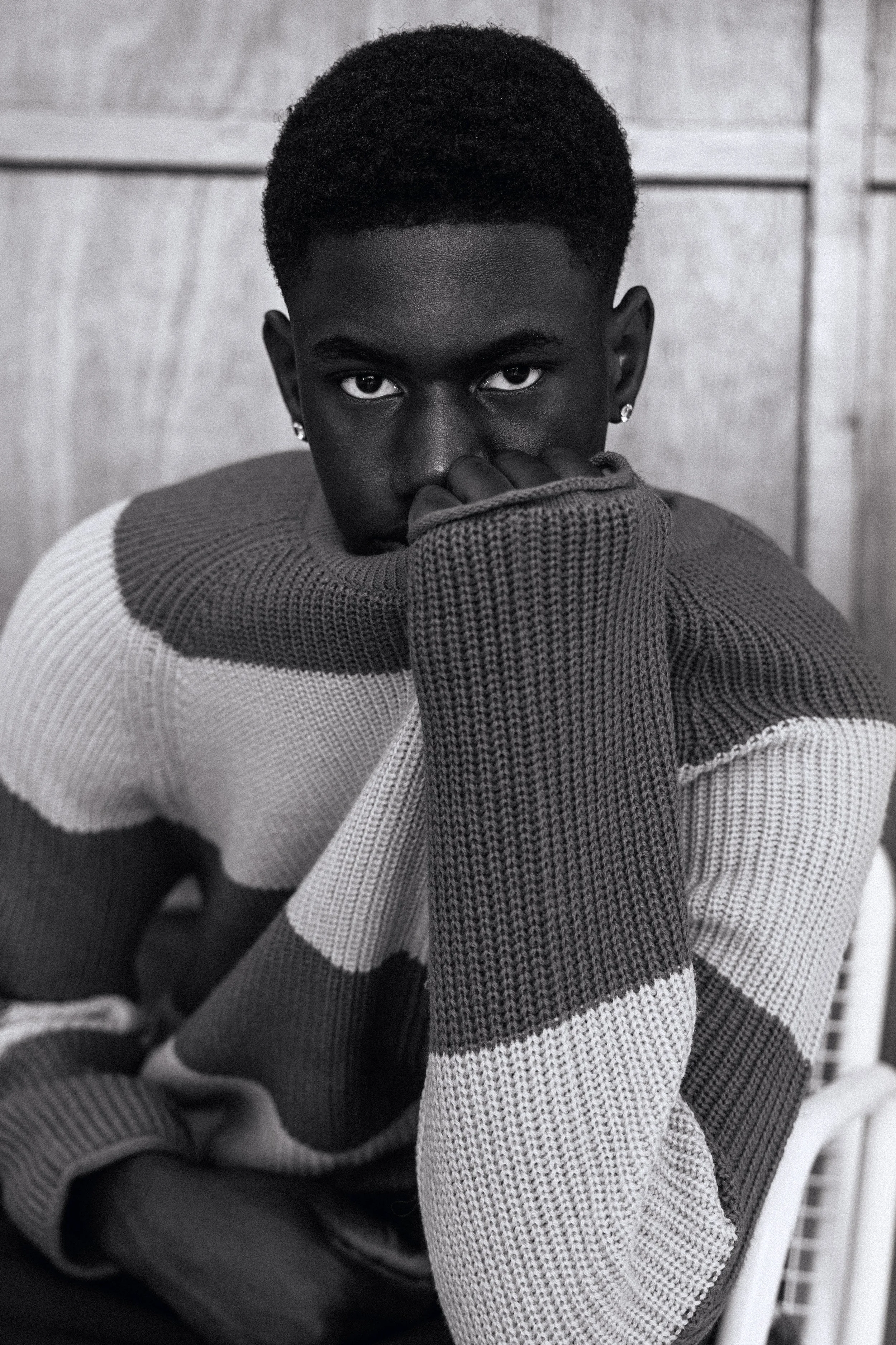 A young Black man with short curly hair looking directly at the camera, resting his chin on his knuckles, wearing earrings and a striped sweater.