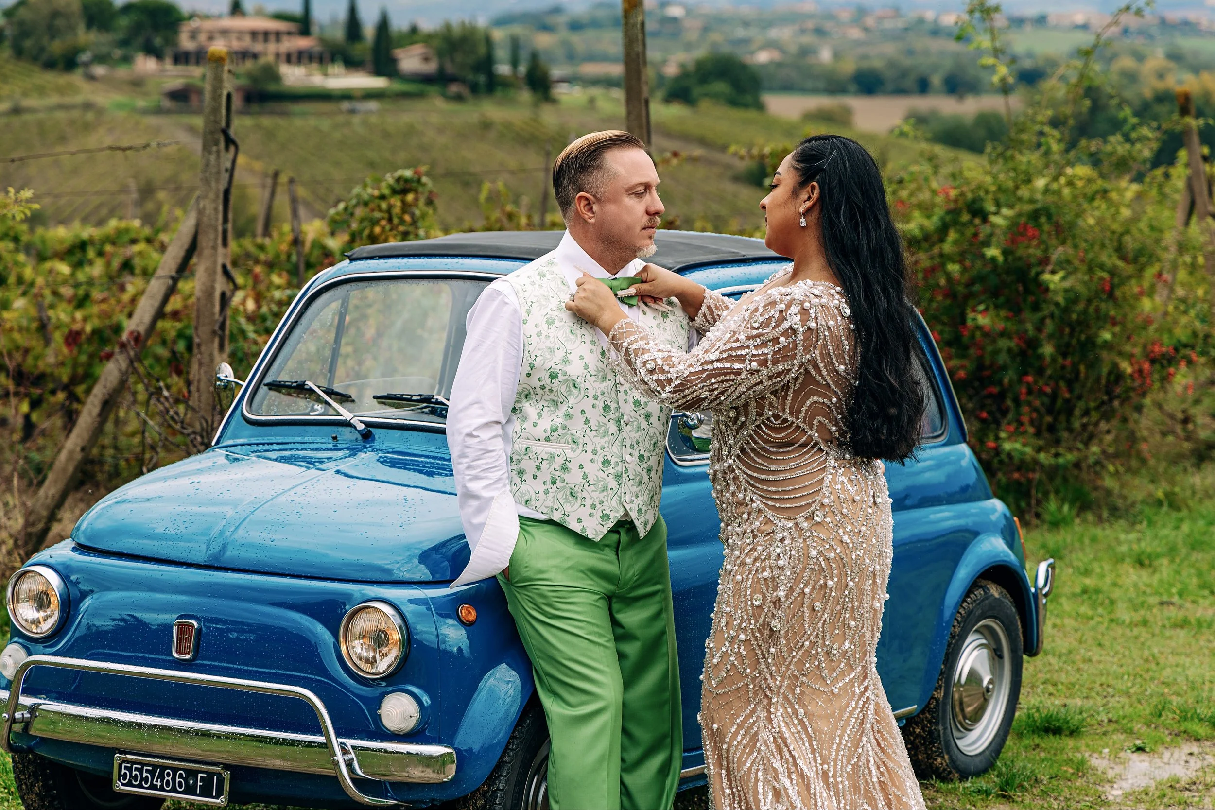 A couple dressed in formal wedding attire standing in front of a blue vintage Fiat car in a scenic countryside vineyard.