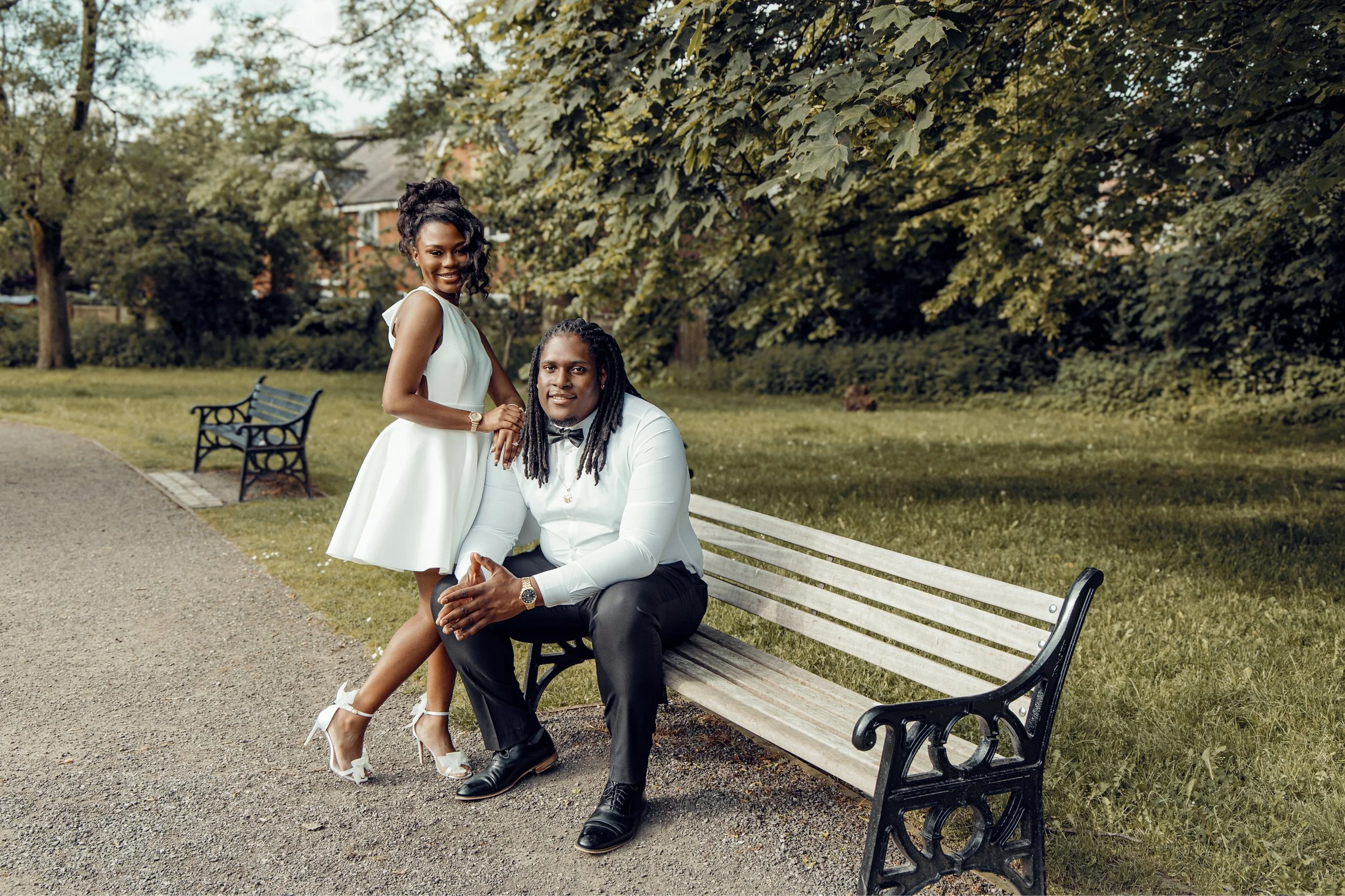 A smiling woman in a white dress and heels standing next to a seated man in a white shirt and black pants, both outdoors in a park with lush trees.