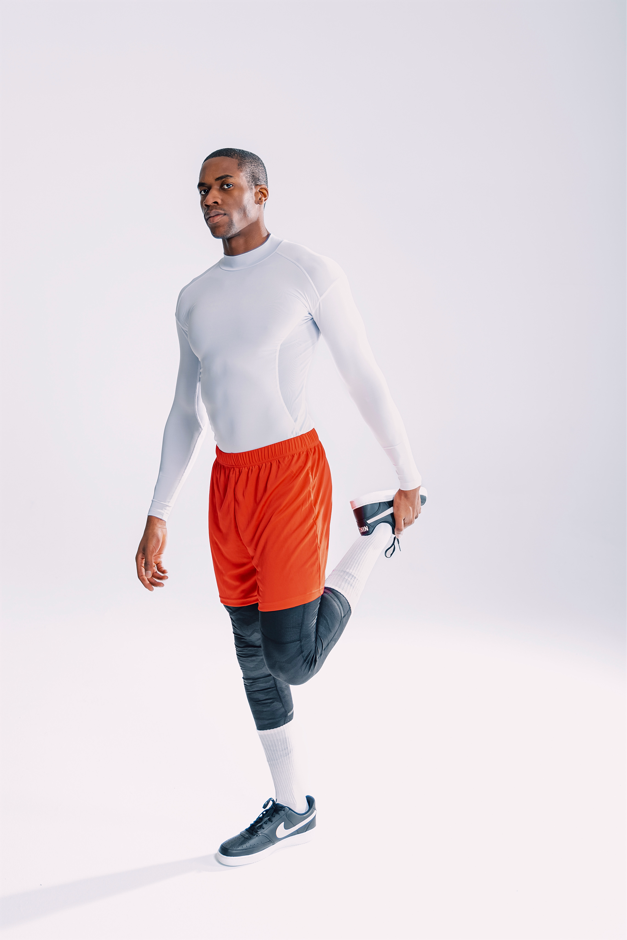 A man in athletic gear stretching his quadriceps in a studio with a plain white background.