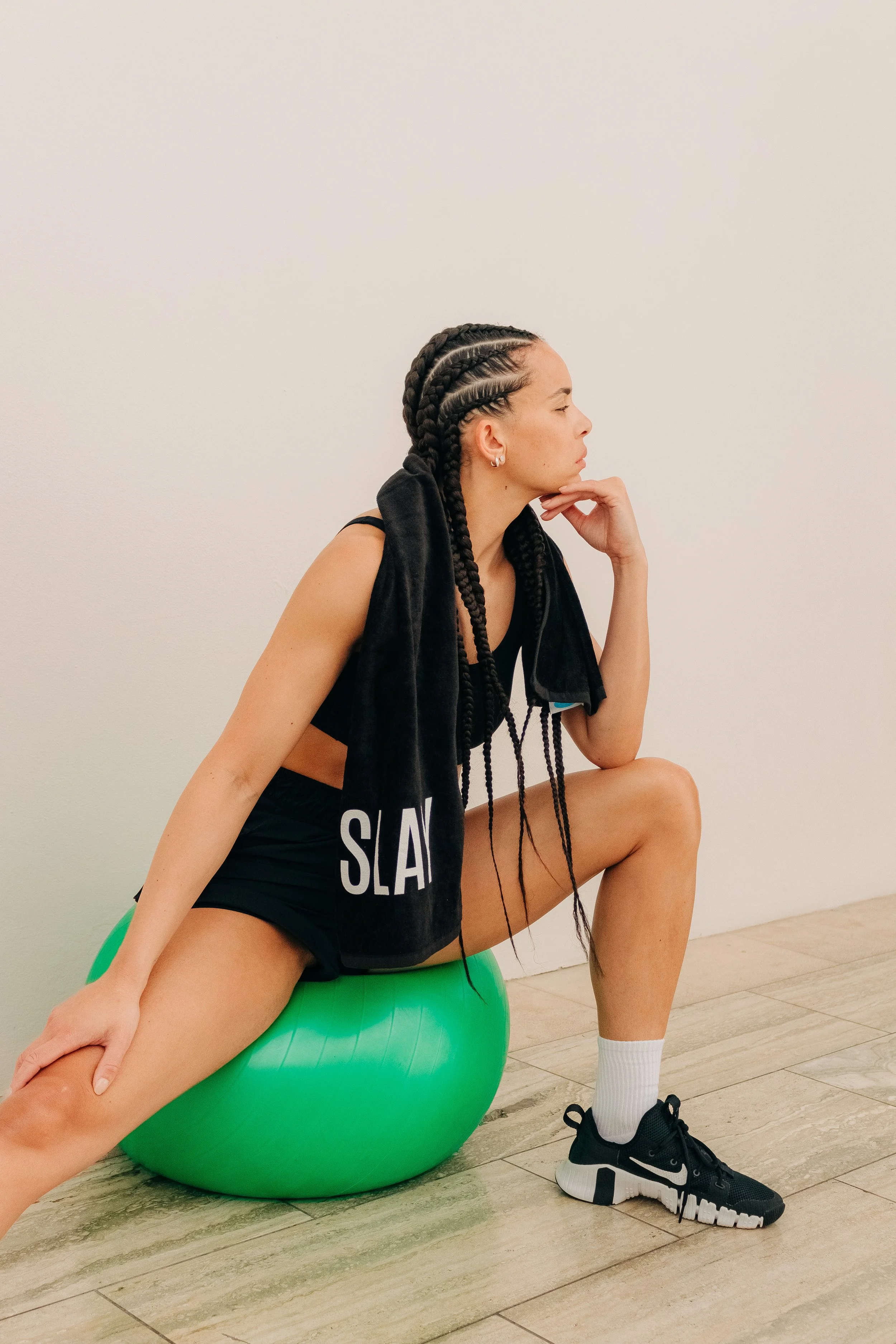 Woman with long braids resting on a green exercise ball in workout clothes, positioned against a plain white wall.