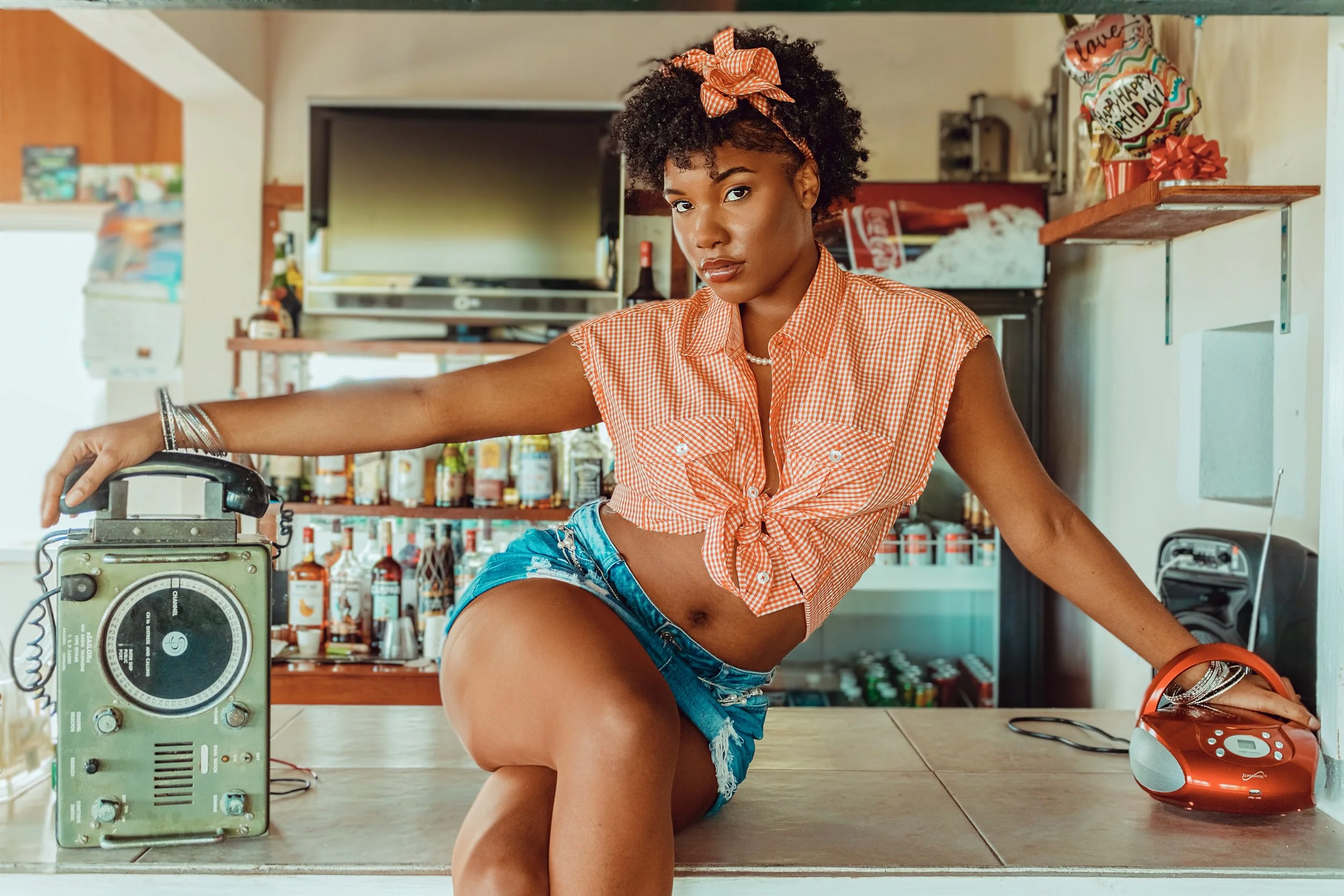 Young woman with curly hair and an orange headband, wearing a tied orange checkered shirt and denim shorts, stretching on a kitchen counter with vintage radio and red portable speaker, with shelves of bottles and a TV in the background.