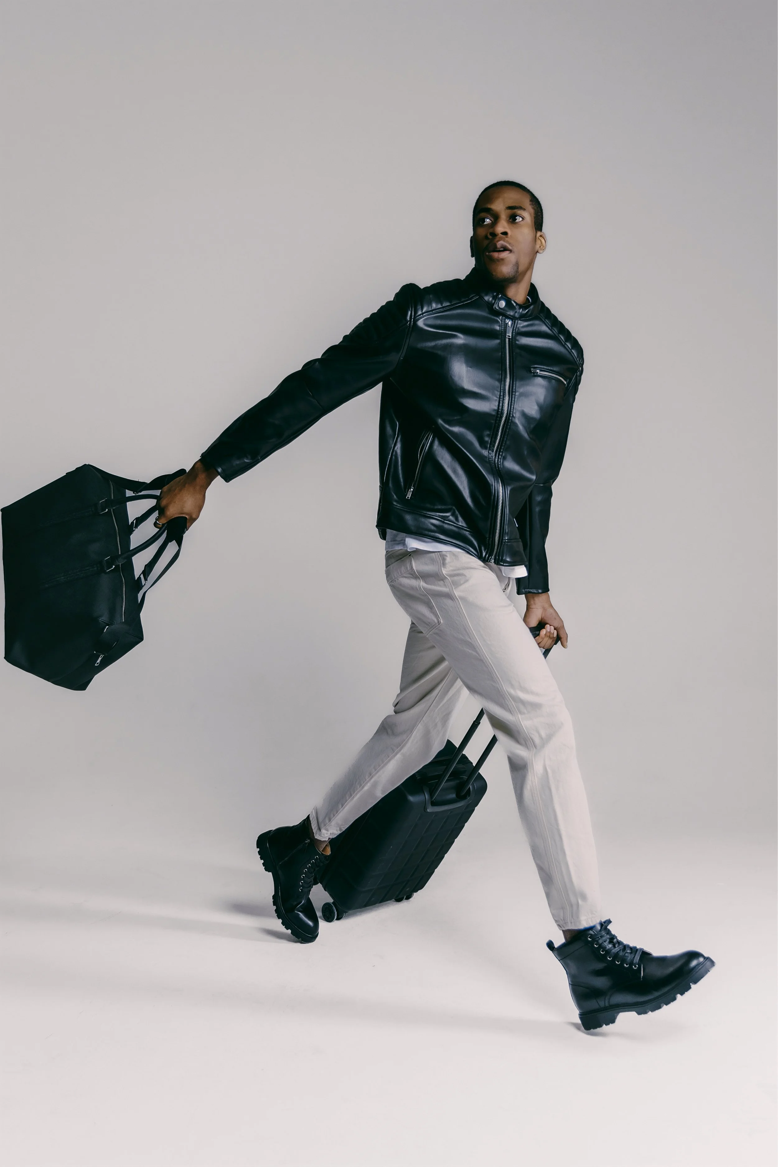 A man dressed in a black leather jacket, beige pants, and black boots walking with rolling luggage in one hand and a black bag in the other against a plain, light-colored background.
