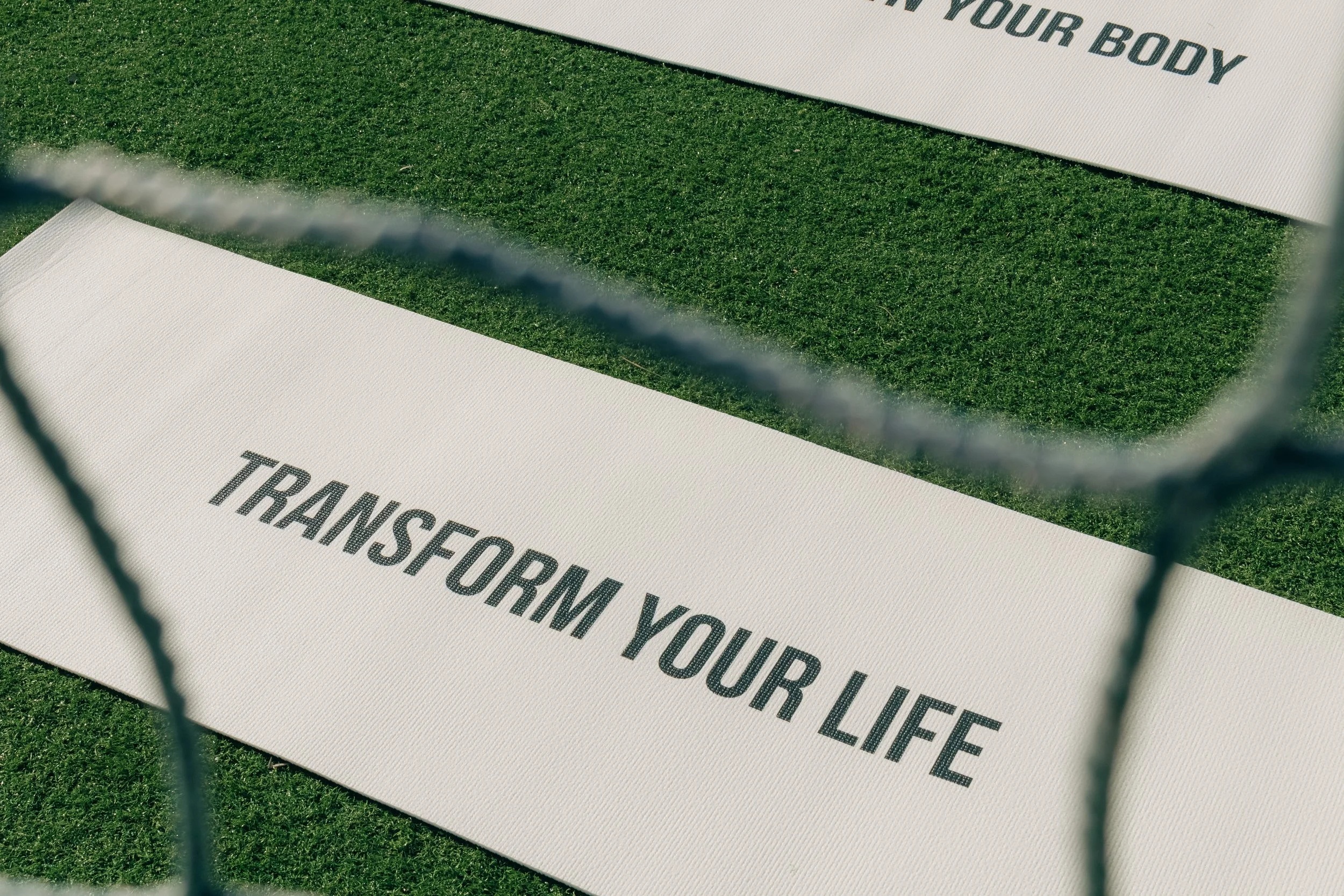 White yoga mats with motivational phrases, one saying 'TRANSFORM YOUR LIFE' and another with 'FEEL YOUR BODY', placed on green grass, viewed through a chain-link fence.