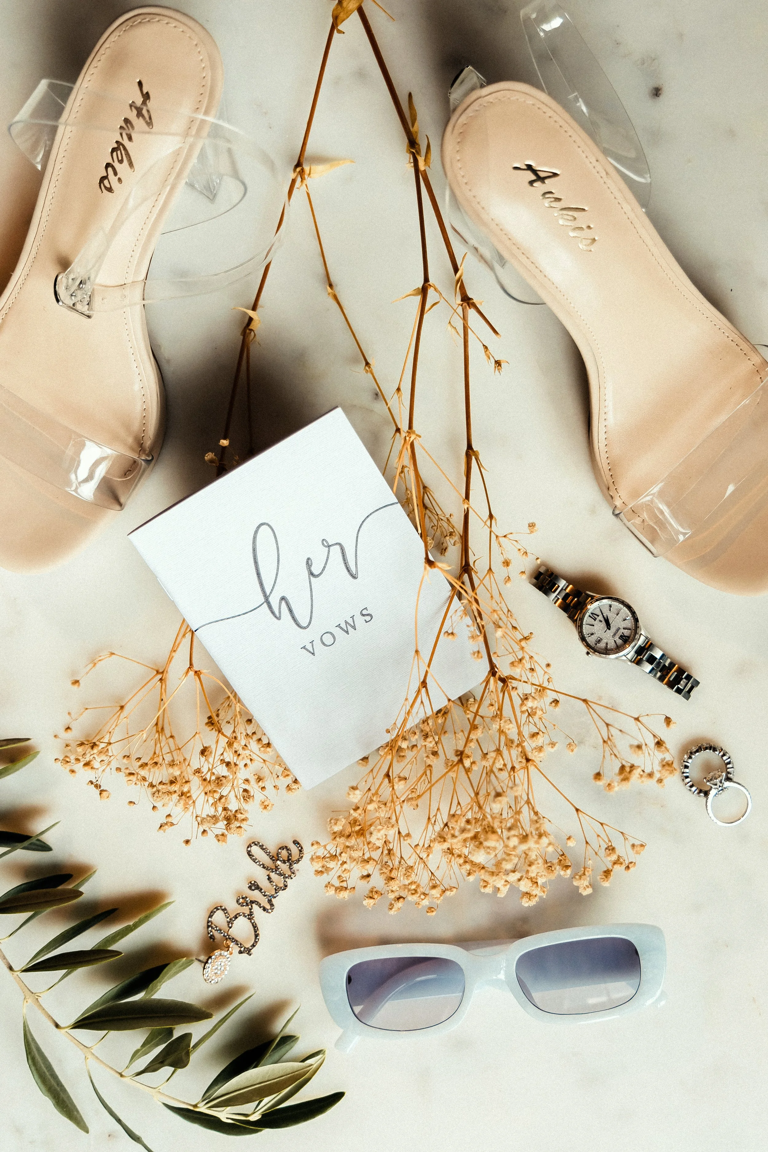 A pair of nude high-heeled shoes with transparent straps, a wristwatch, a ring, a pair of white sunglasses, a greeting card, dried flowers, and some greenery arranged on a white surface.