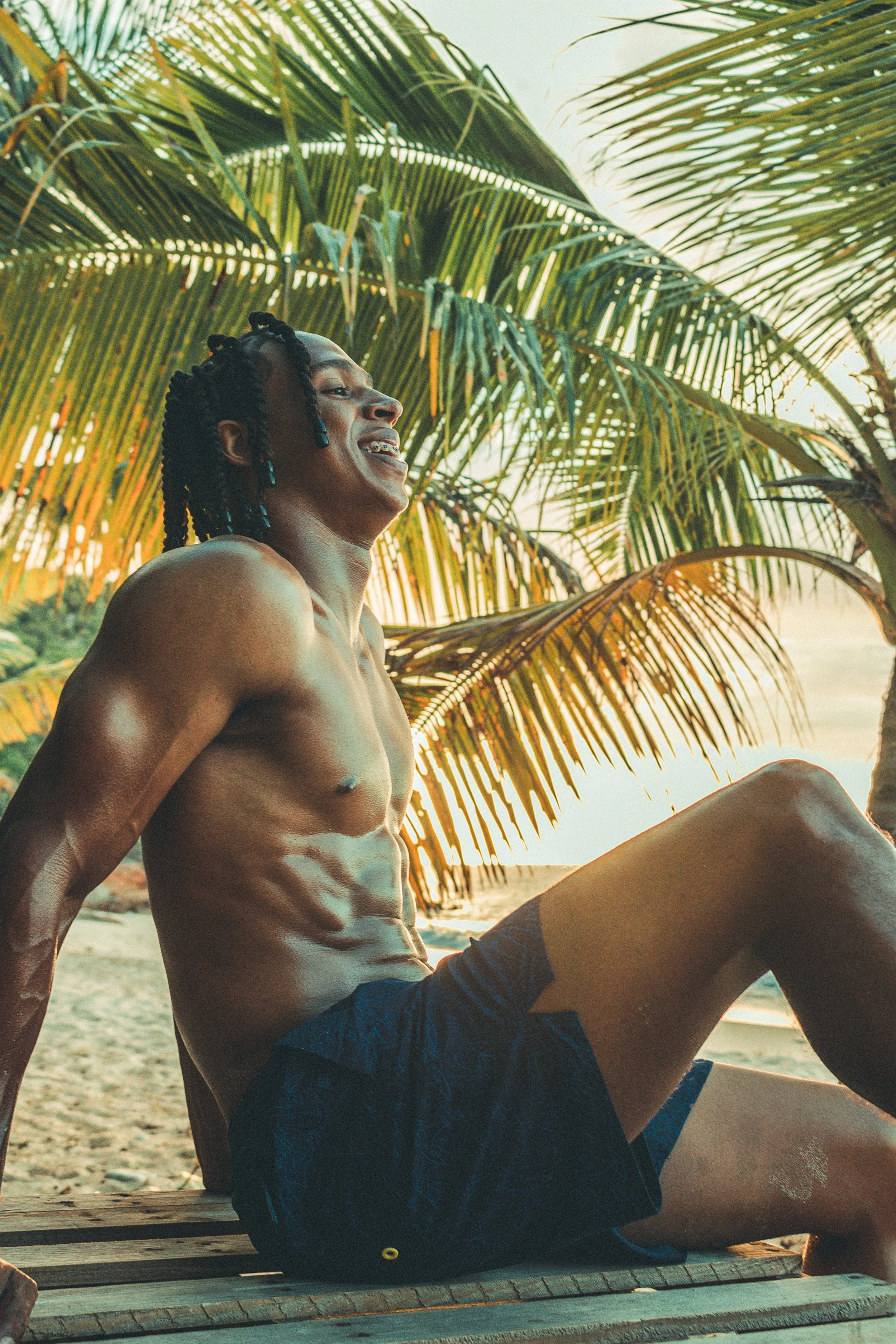 A shirtless man with dreadlocks smiling and sitting outdoors under palm trees, wearing shorts, with a sunset or sunrise in the background.