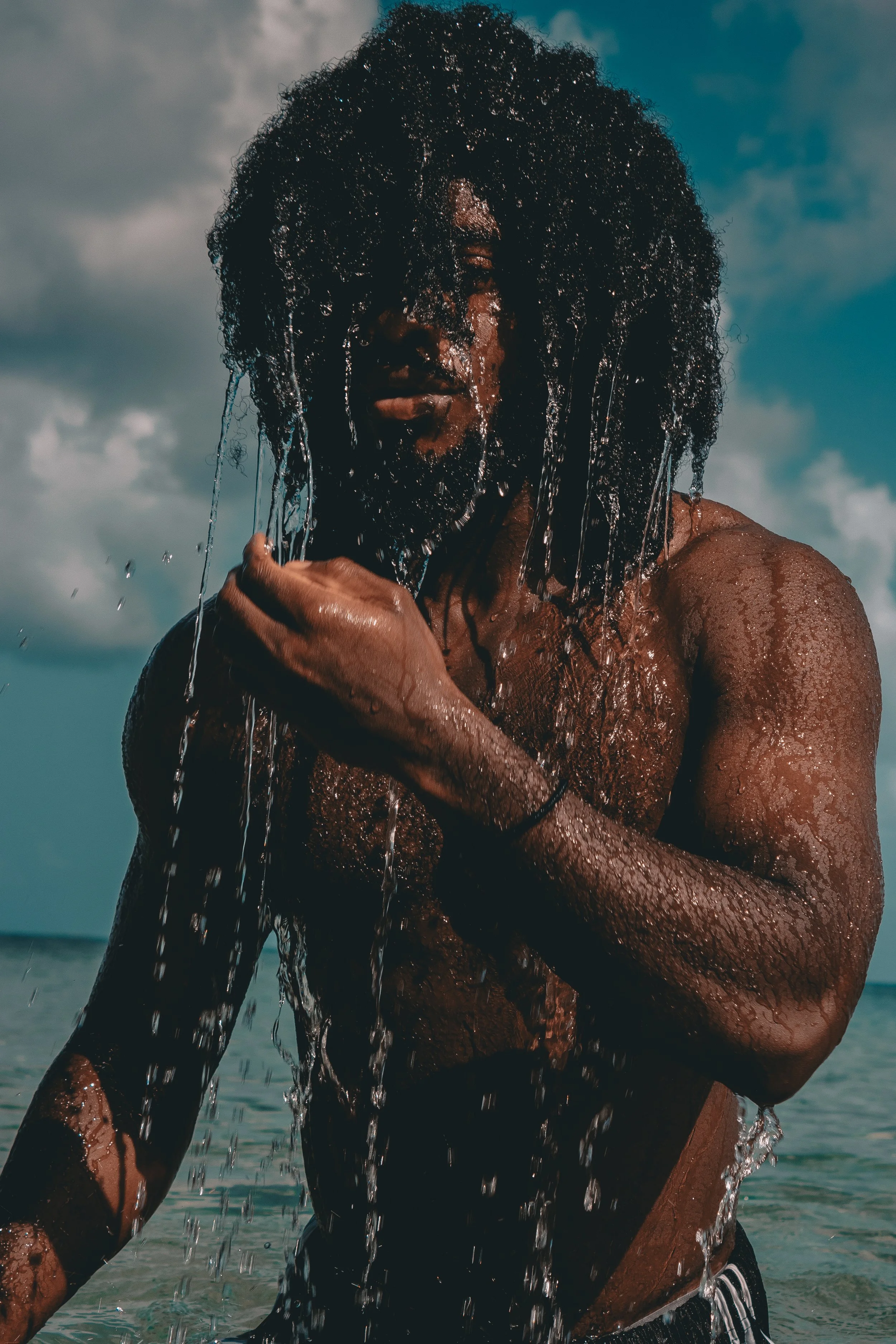 A person with dark, curly hair is at the beach, pouring water over their face and upper body, with the ocean and cloudy sky in the background.
