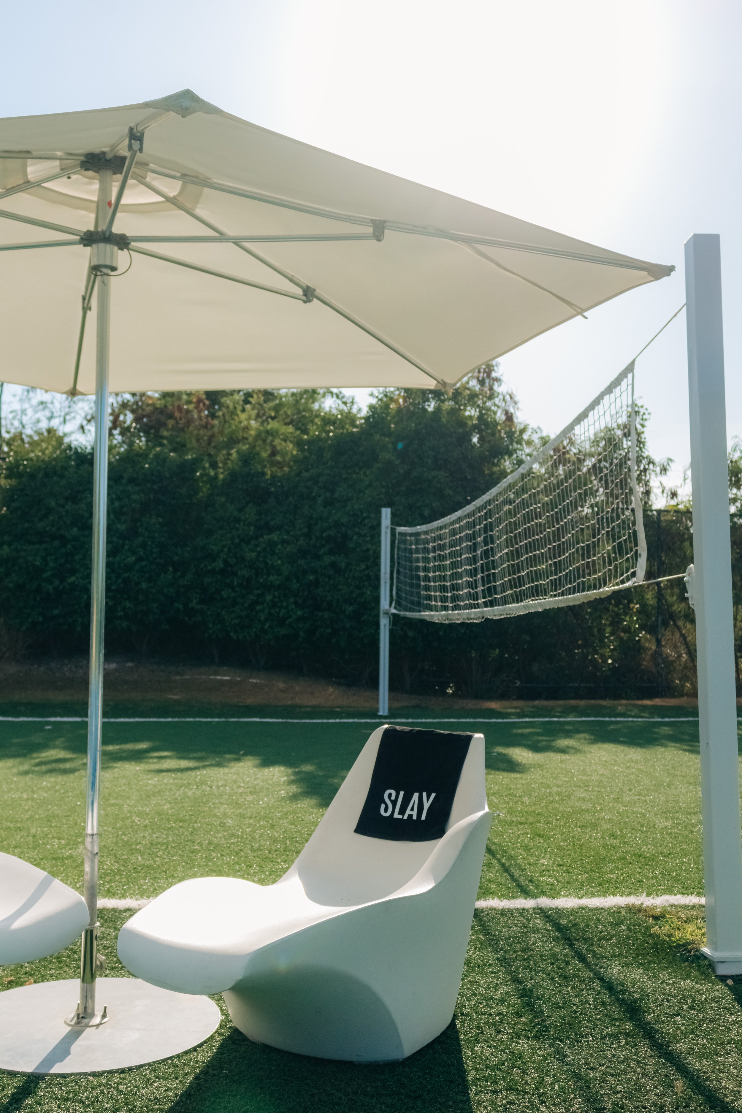 Beachside lounge chair with a black towel that says 'SLAY', under a large white umbrella, near a volleyball net on green artificial turf.