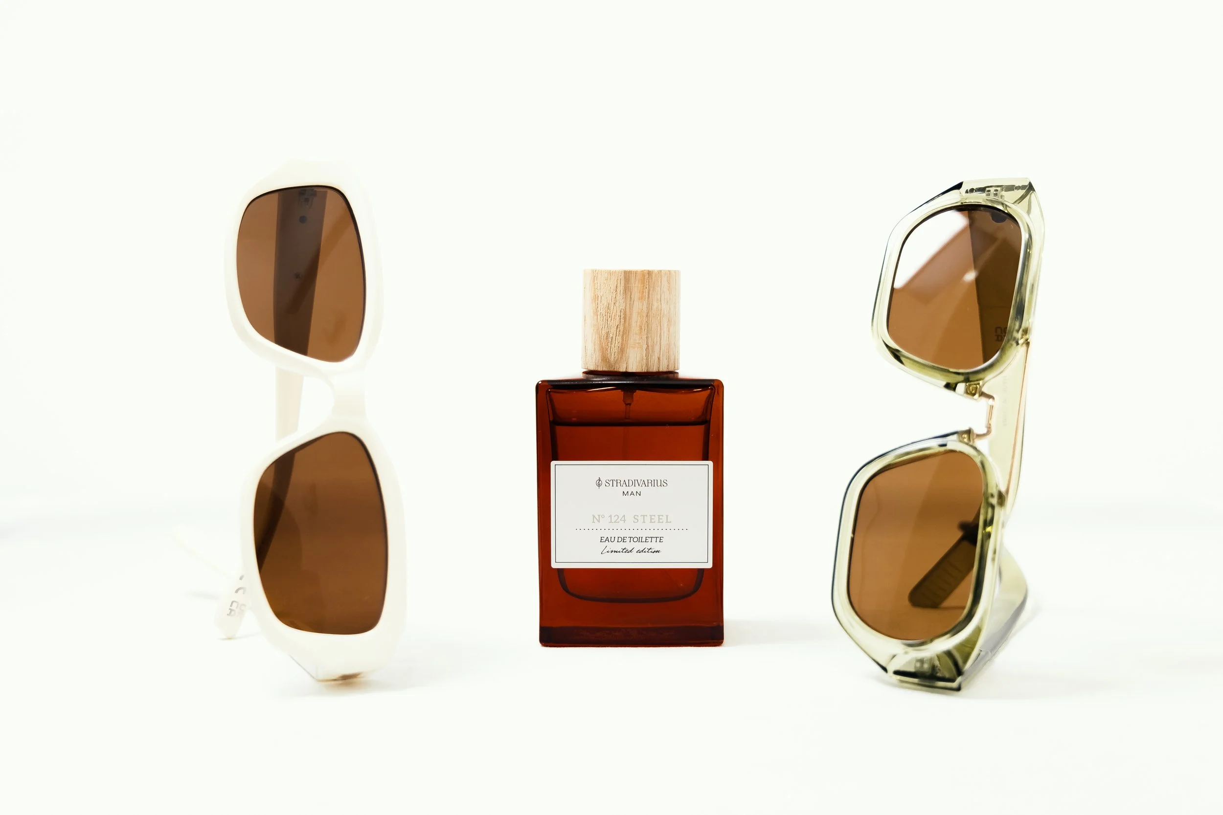 Pair of white sunglasses on the left, amber-colored perfume bottle with a wooden cap in the center, and clear sunglasses with amber lenses on the right, all against a plain white background.