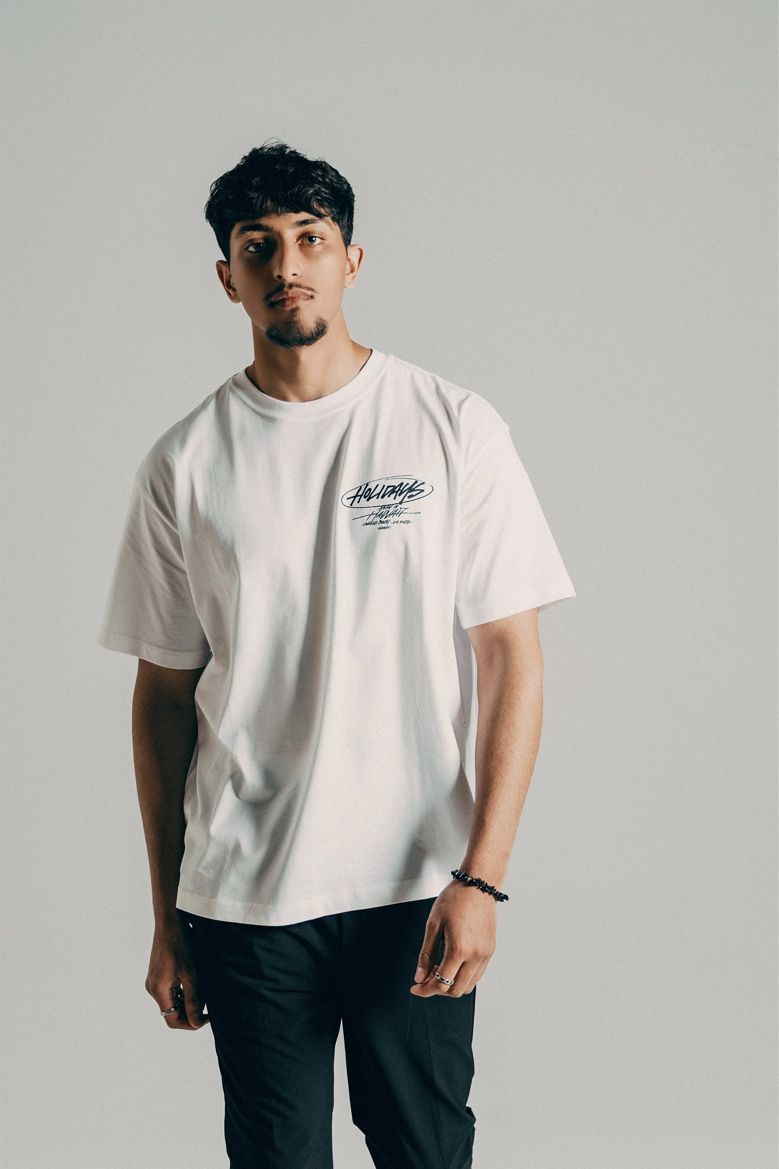 A young man with dark hair and a goatee, wearing a white t-shirt with a graphic logo on the chest, standing against a plain light gray background.