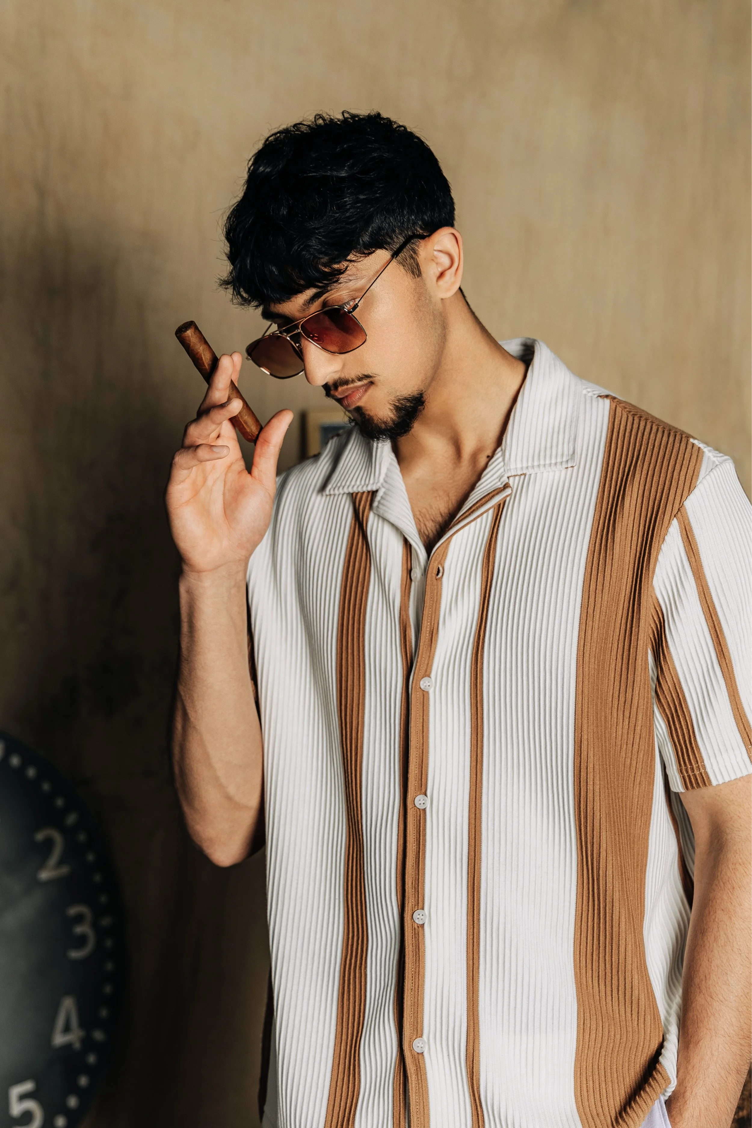A man with black hair, sunglasses, and facial hair holds a cigar to his ear, standing in front of a beige wall and a large clock.