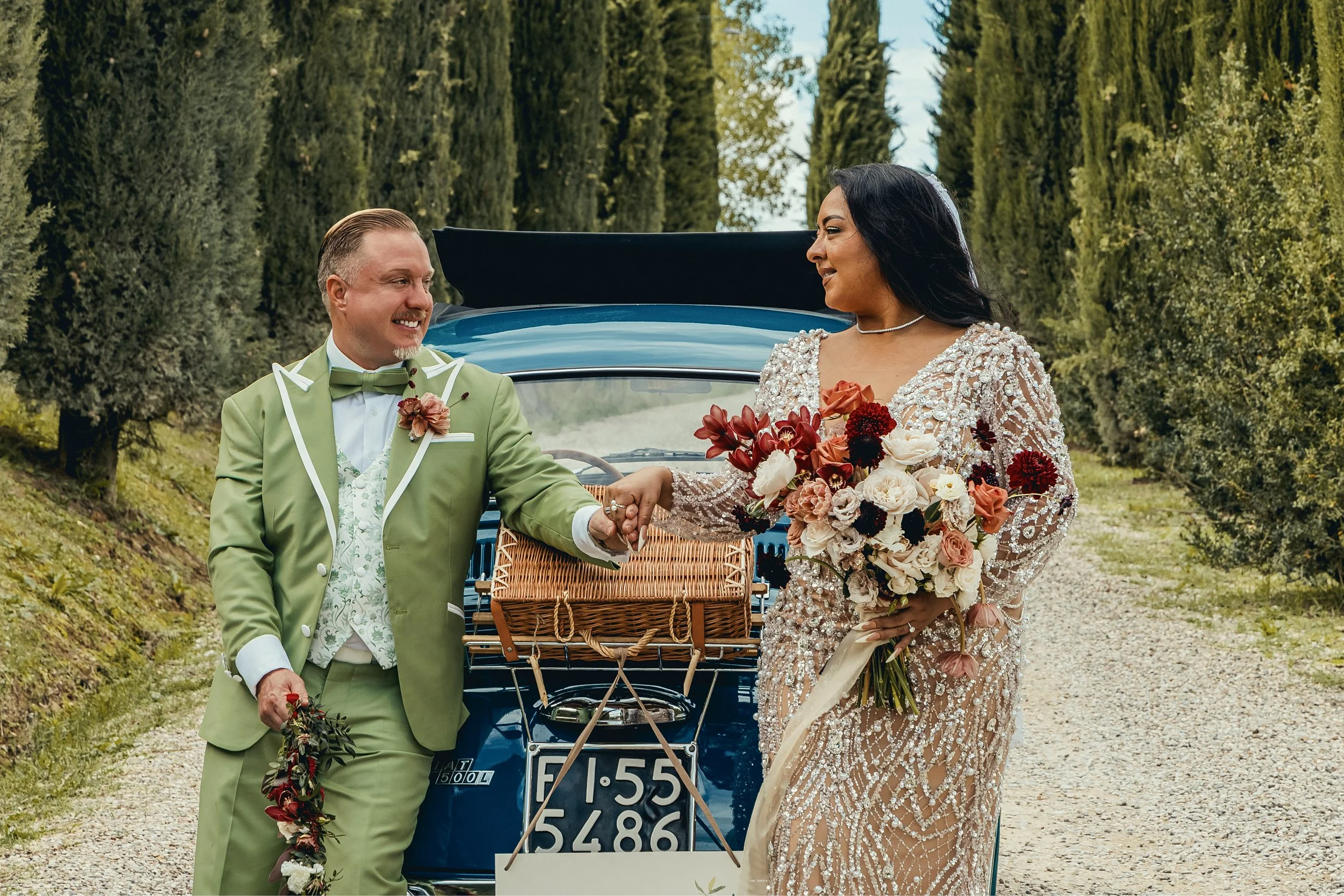 A couple dressed in formal wedding attire smiling and holding hands outdoors in front of a vintage blue car surrounded by tall green trees.