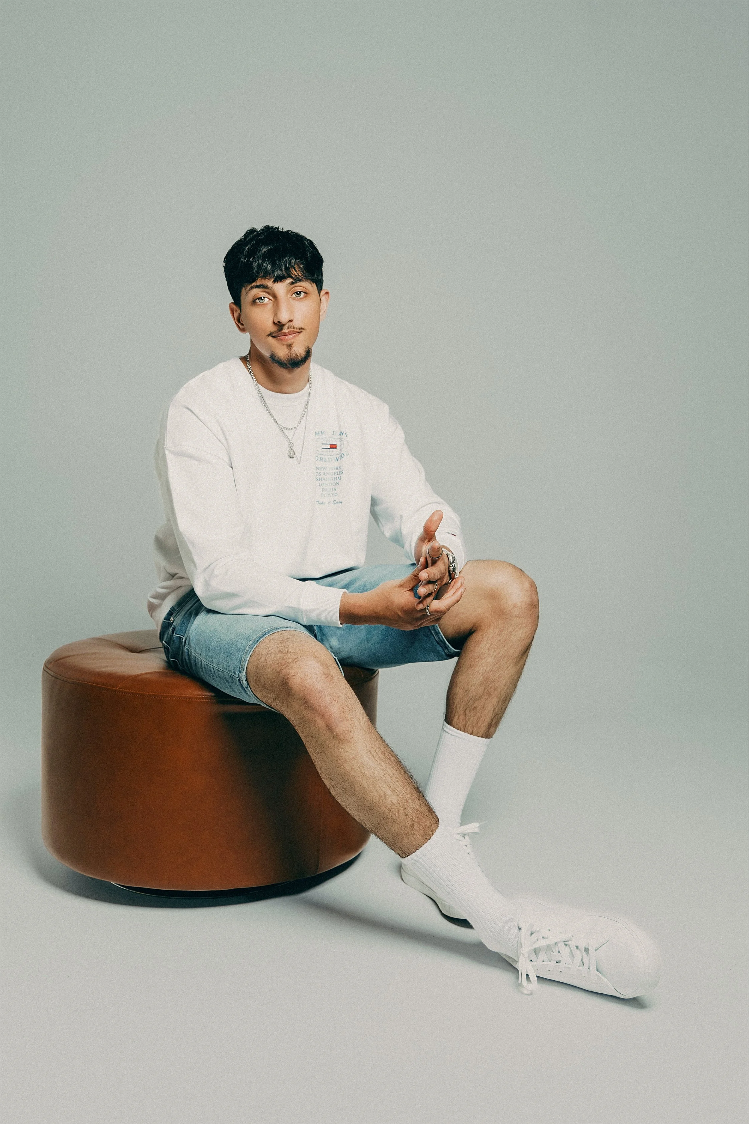 A young man sitting on a round leather ottoman, wearing a white sweatshirt, denim shorts, white socks, and sneakers, against a plain background.