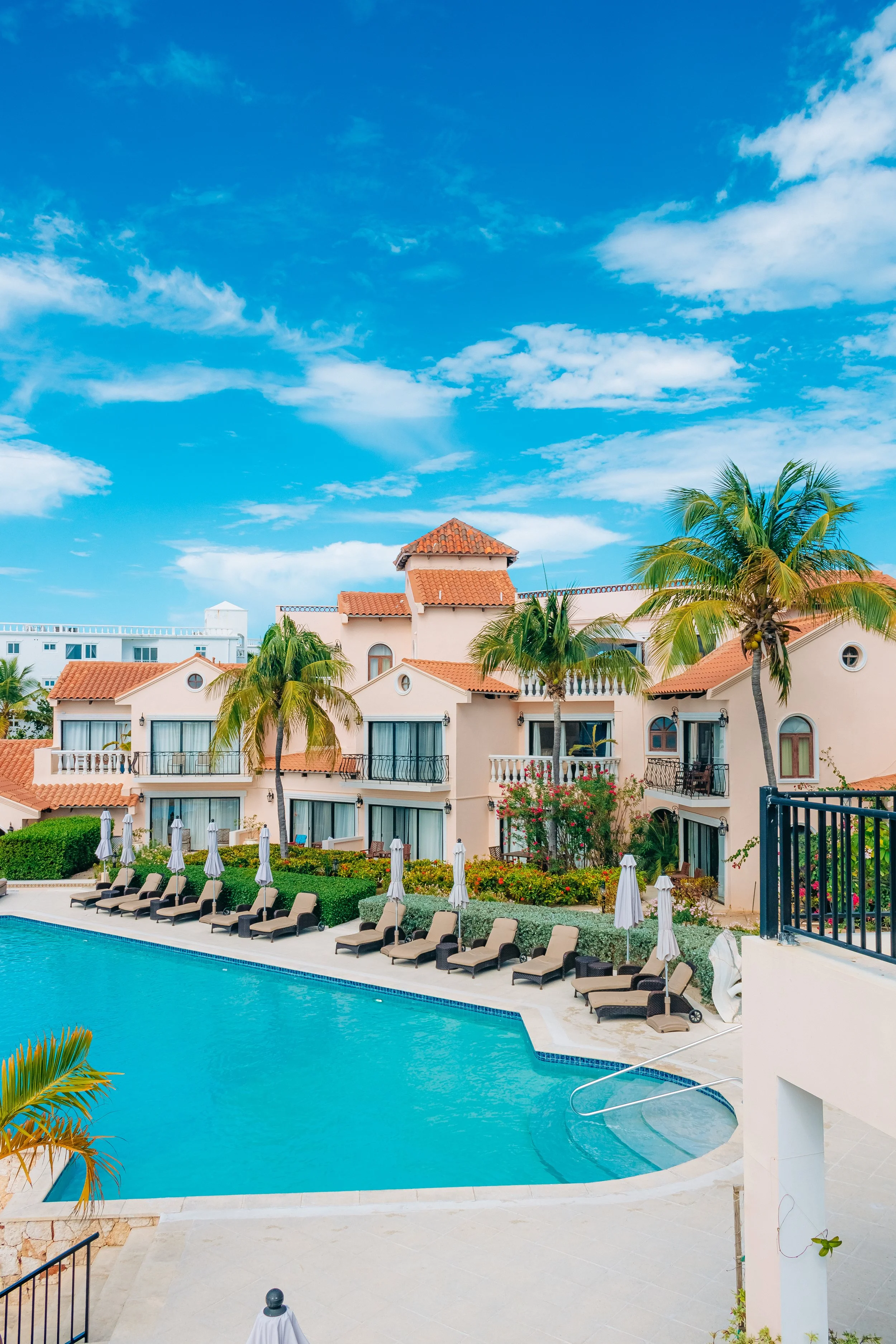 A view of a luxurious hotel pool area with lounge chairs, umbrellas, and lush palm trees, with a building featuring terracotta roof tiles and a clear blue sky with scattered clouds in the background.