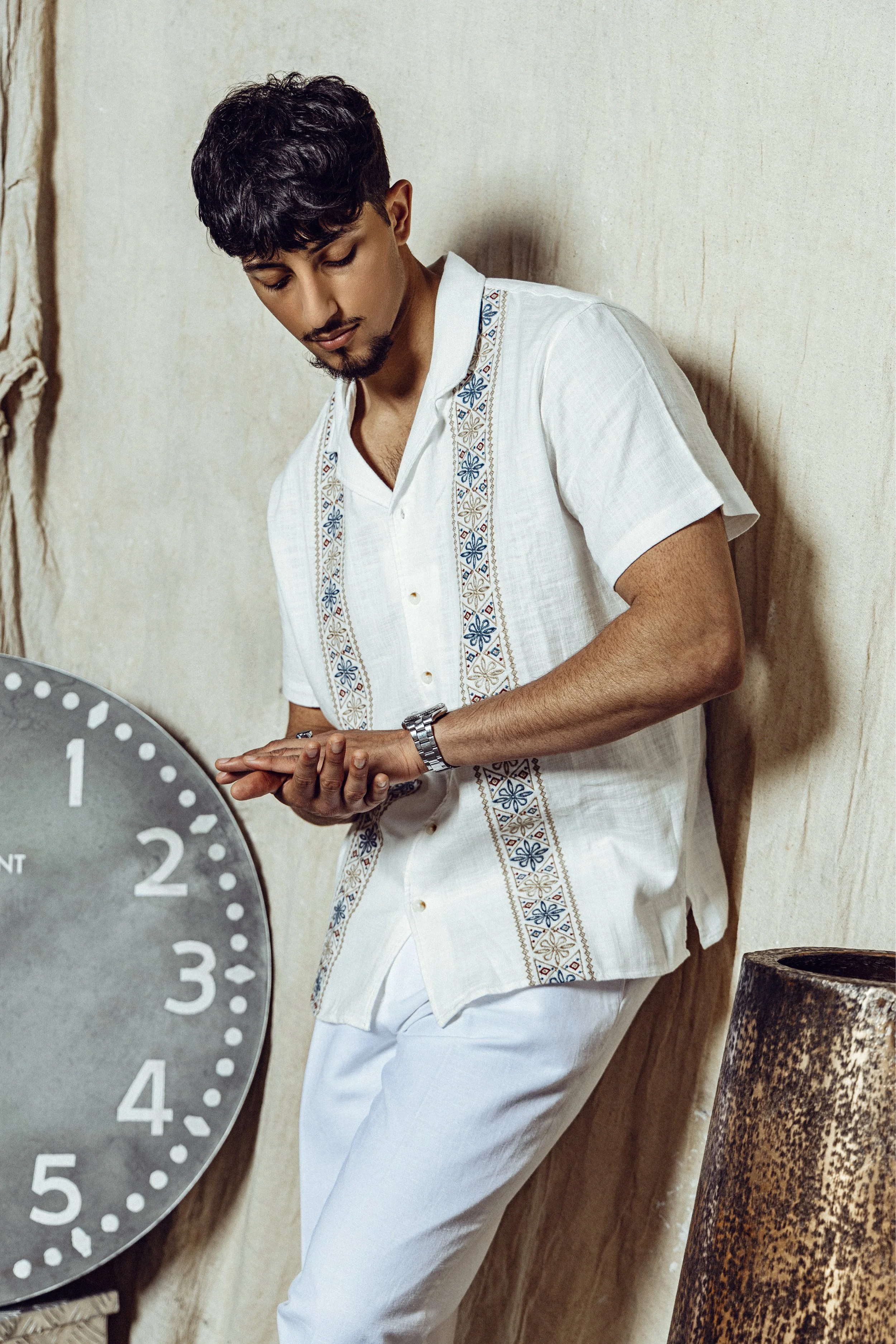A young man with dark hair and a goatee, dressed in a white shirt with embroidered blue and brown patterns, stands against a beige textured wall, looking at his watch while leaning on a wall. A large gray clock with white numbers and a rusted cylindr
