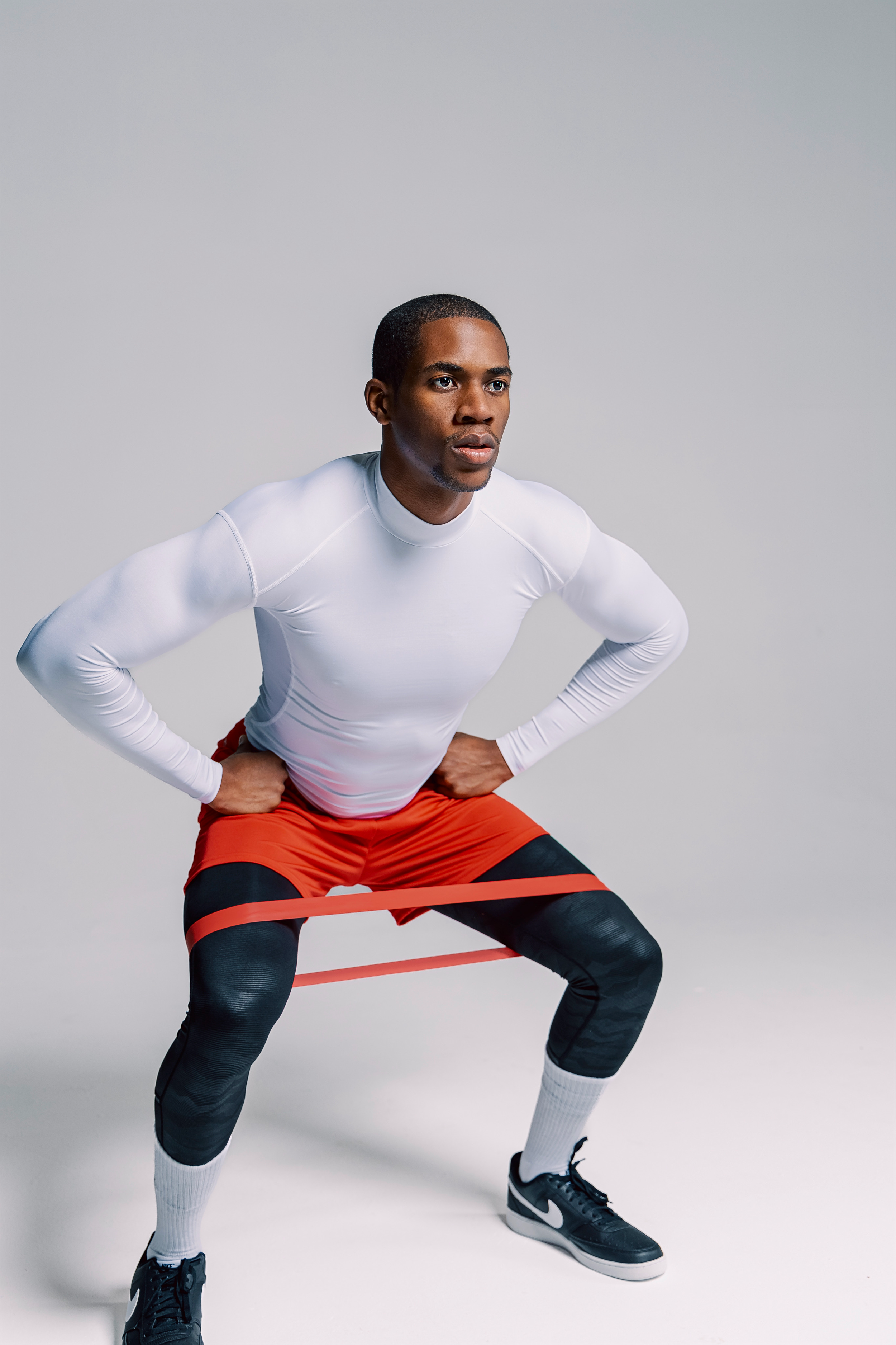 A man in athletic clothing, including a white long-sleeve shirt, red shorts, black leggings, white socks, and black Nike sneakers, is exercising with resistance bands around his thighs in a squat position.