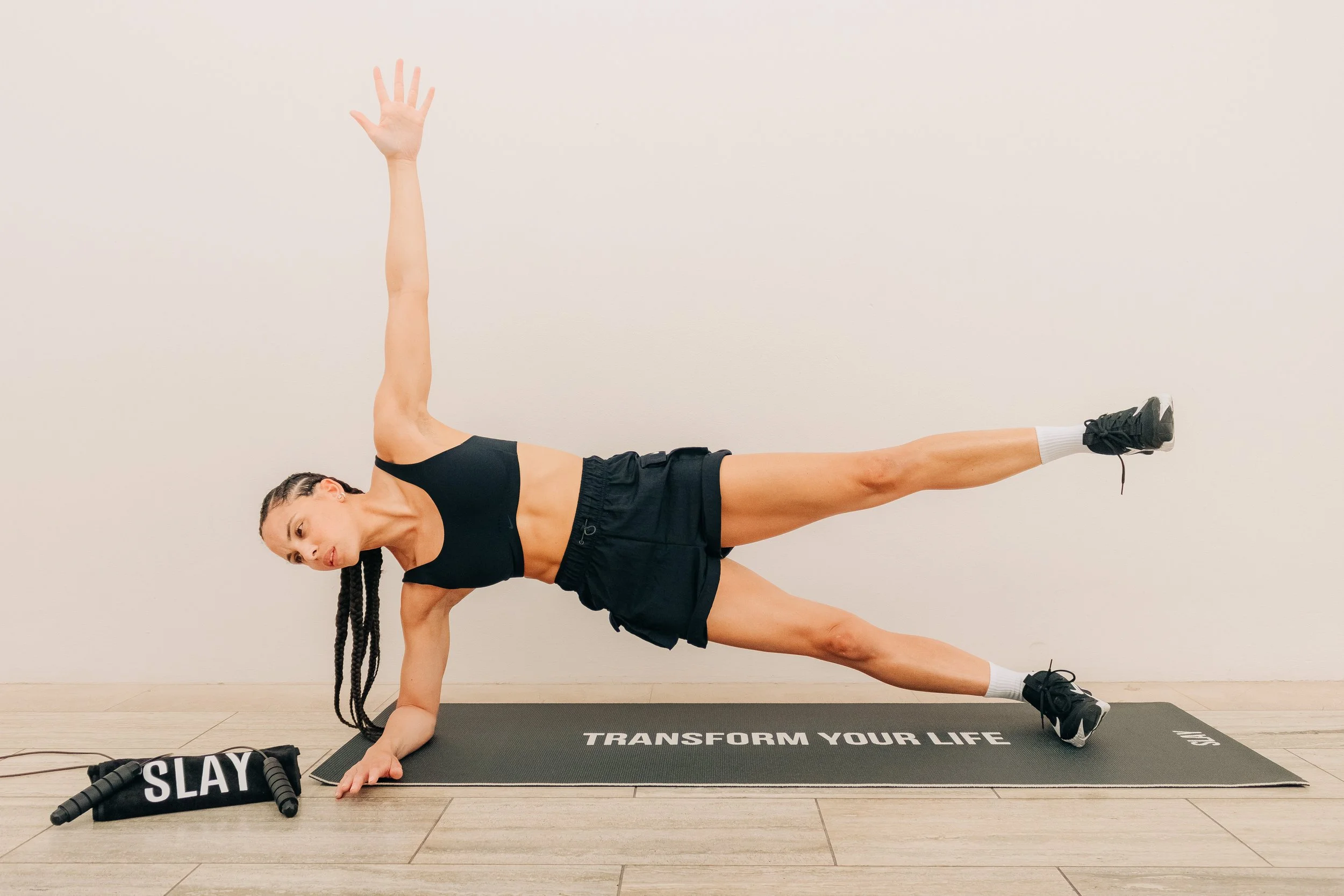 Woman performing a side plank exercise on a mat with the words 'Transform Your Life' printed on it, in a room with wooden flooring and a plain wall.