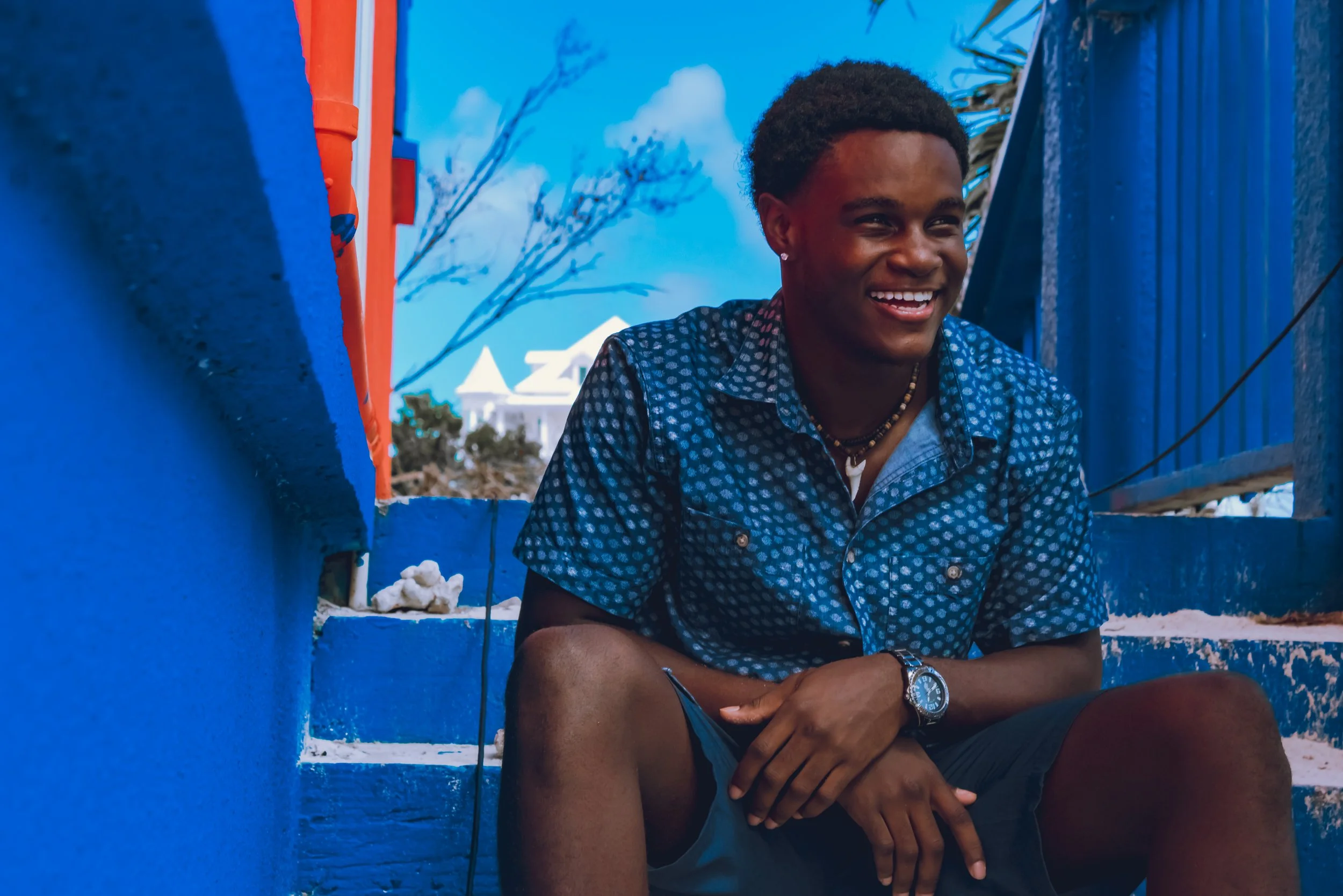 A young man with dark skin and short curly hair smiling while sitting outdoors against a blue fence with a bright sky and trees in the background. He is wearing a blue patterned shirt, a beaded necklace, earrings, a watch, and has one arm resting on 