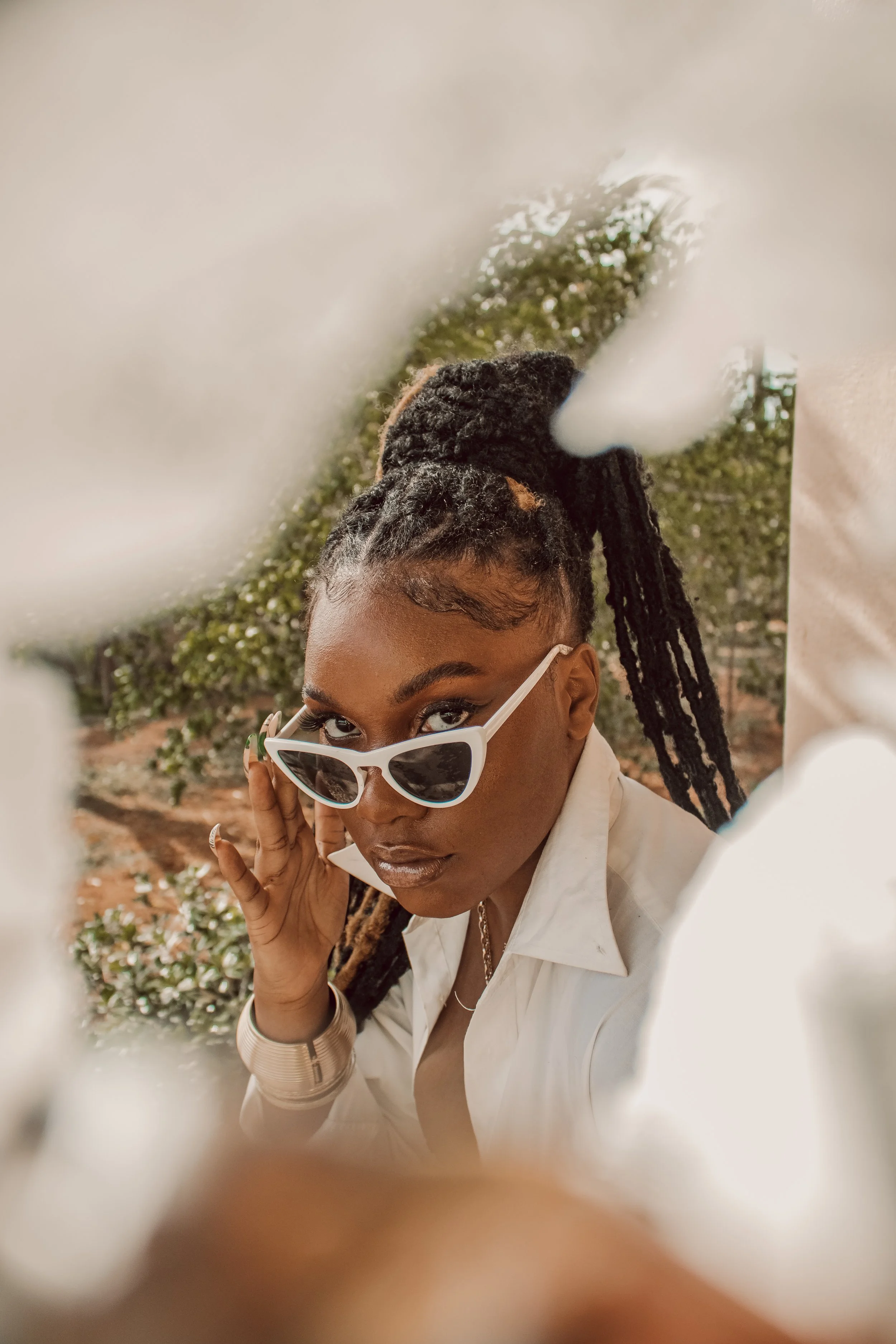A woman with braided hair and dark skin, wearing white sunglasses and a white shirt, looks over her shoulder through an opening. She is outdoors with greenery in the background.
