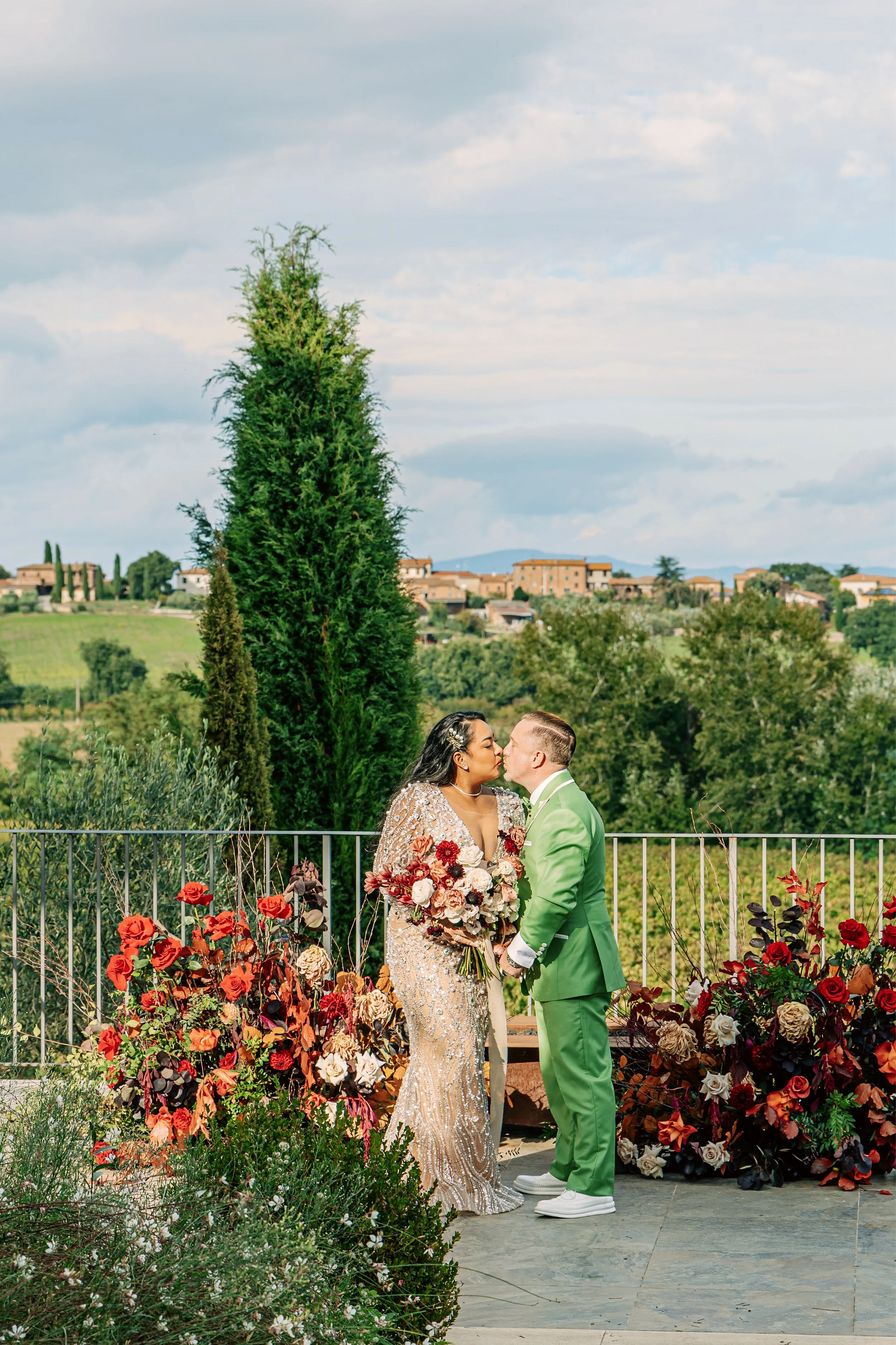 A bride and groom sharing a kiss outdoors during their wedding ceremony, with colorful flowers and a scenic landscape in the background.