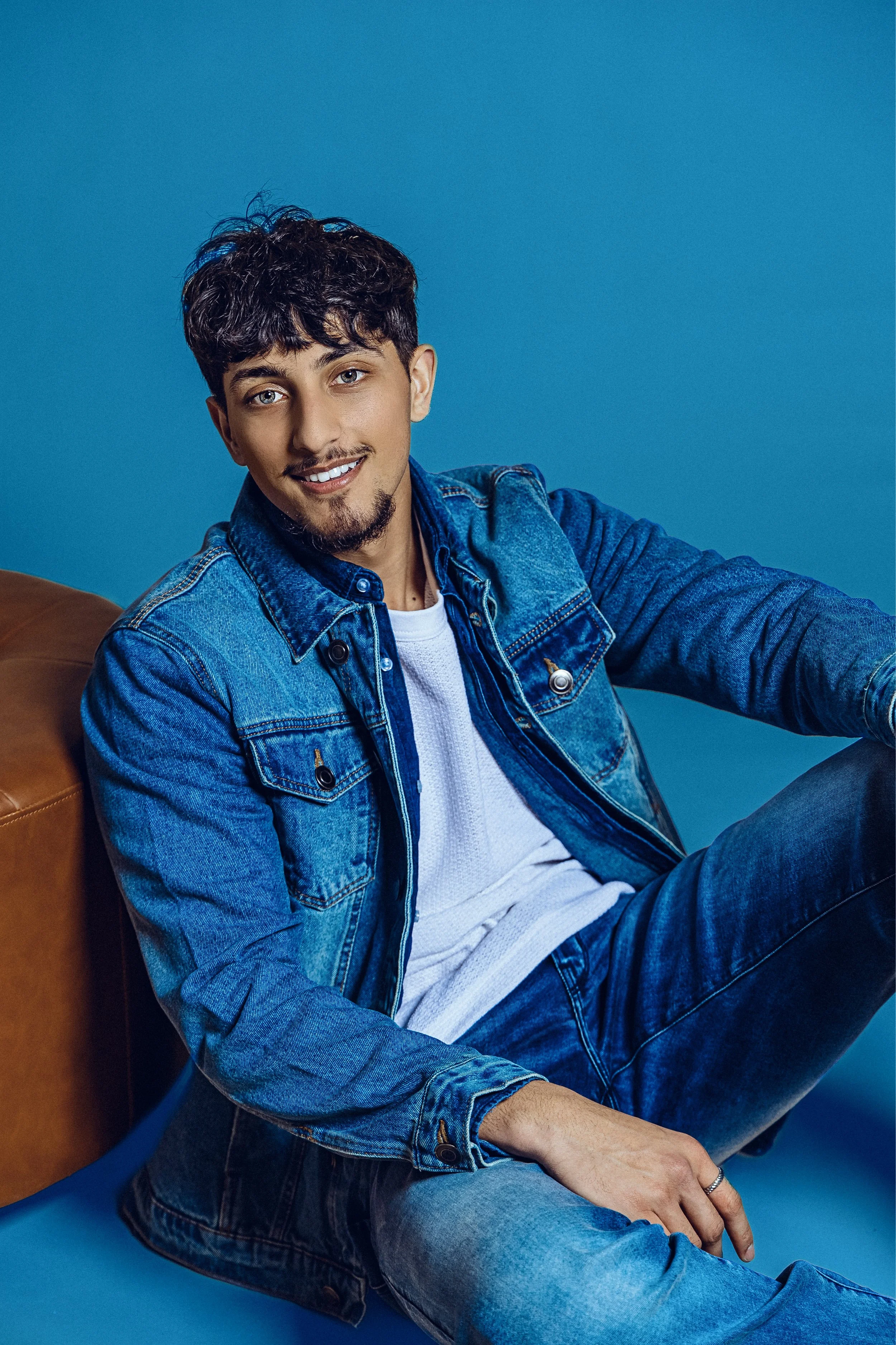 A young man with dark, curly hair and a light beard, wearing a denim jacket over a white shirt, sitting and leaning against a brown leather chair on a blue background, smiling at the camera.