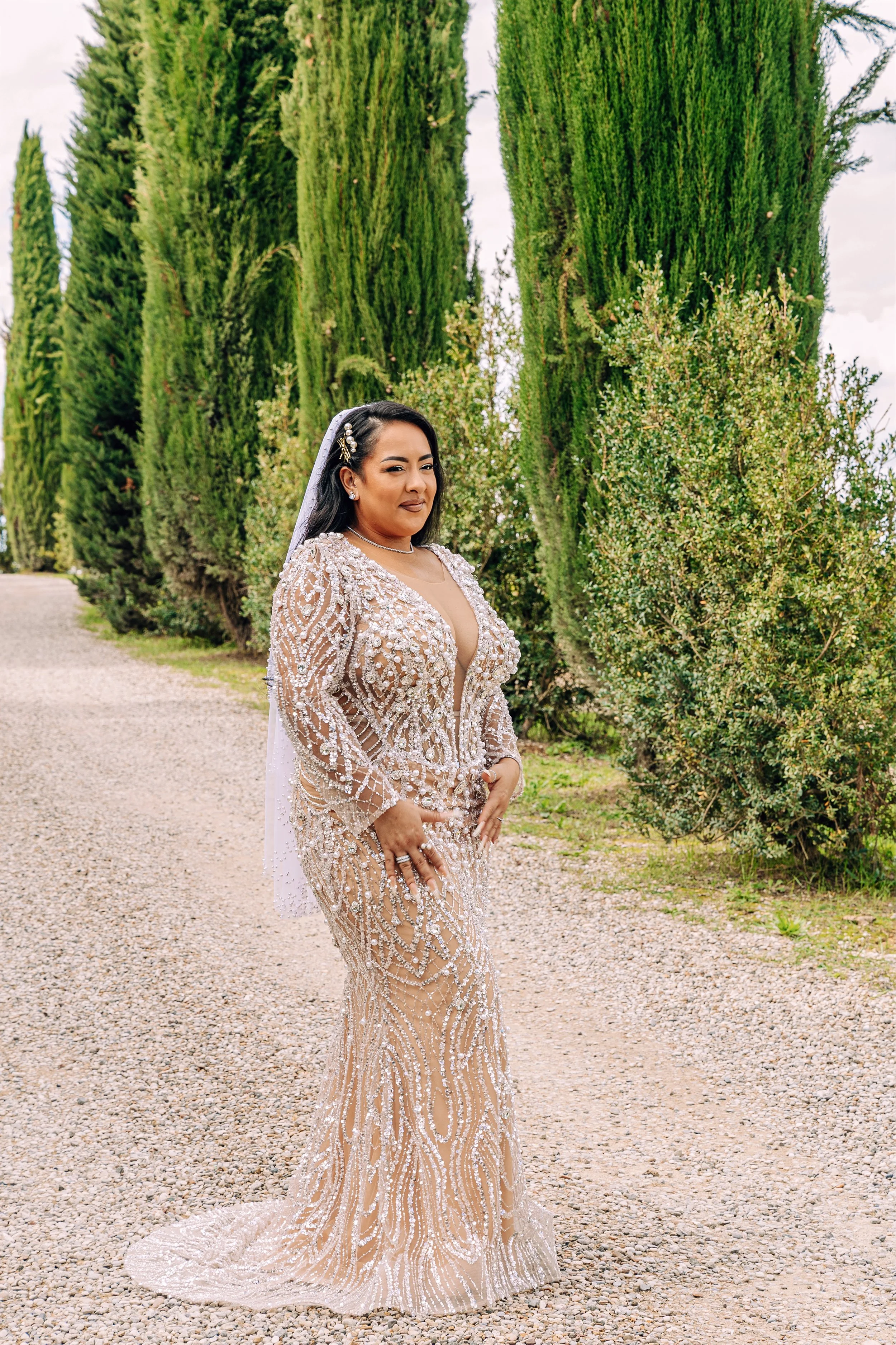 A woman in a detailed, beaded gown with a veil, standing outdoors on a gravel path with tall green trees in the background.