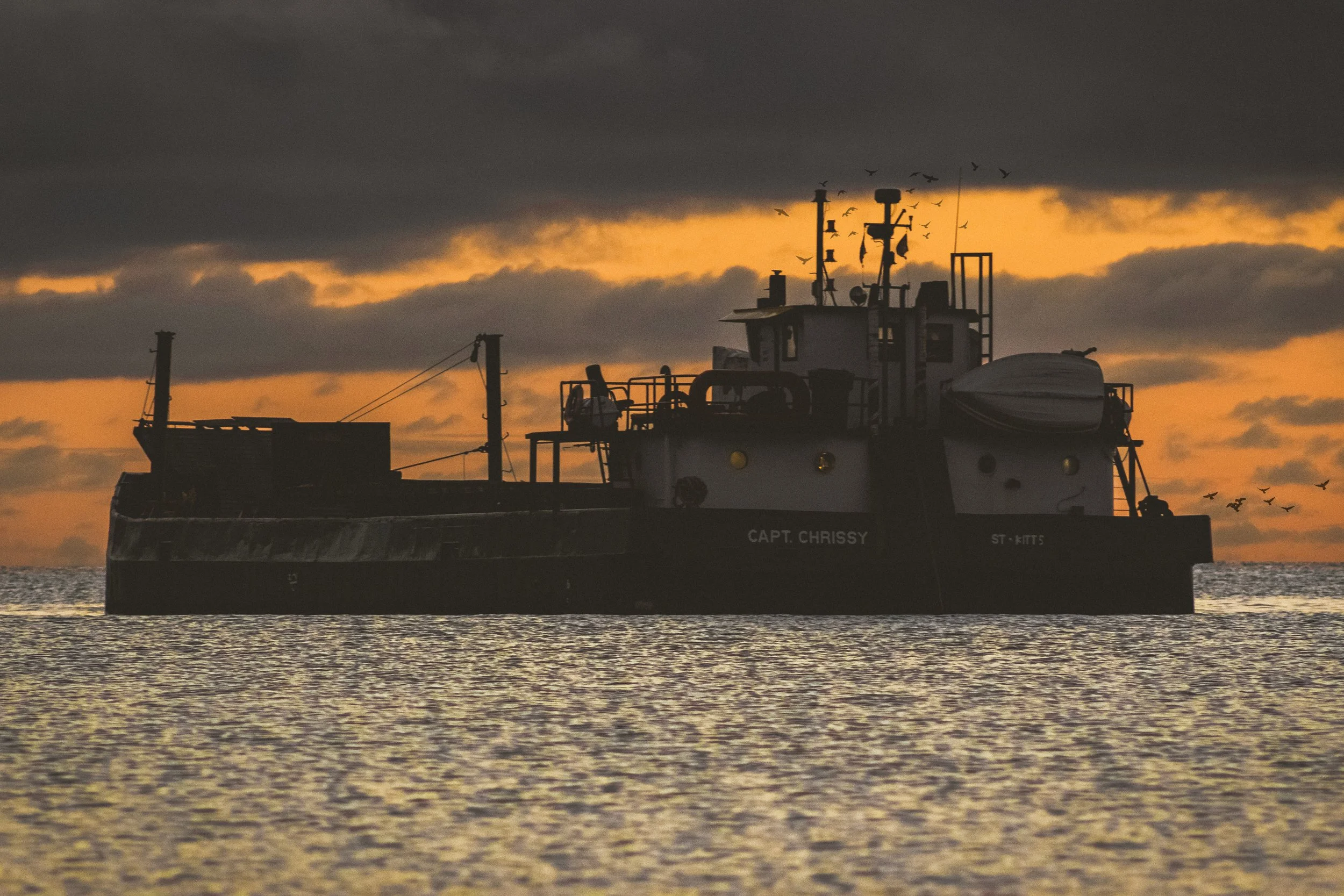 Silhouette of a boat labeled 'Capt. Chrissy' on the water at sunset, with birds flying in the sky and dark clouds overhead.