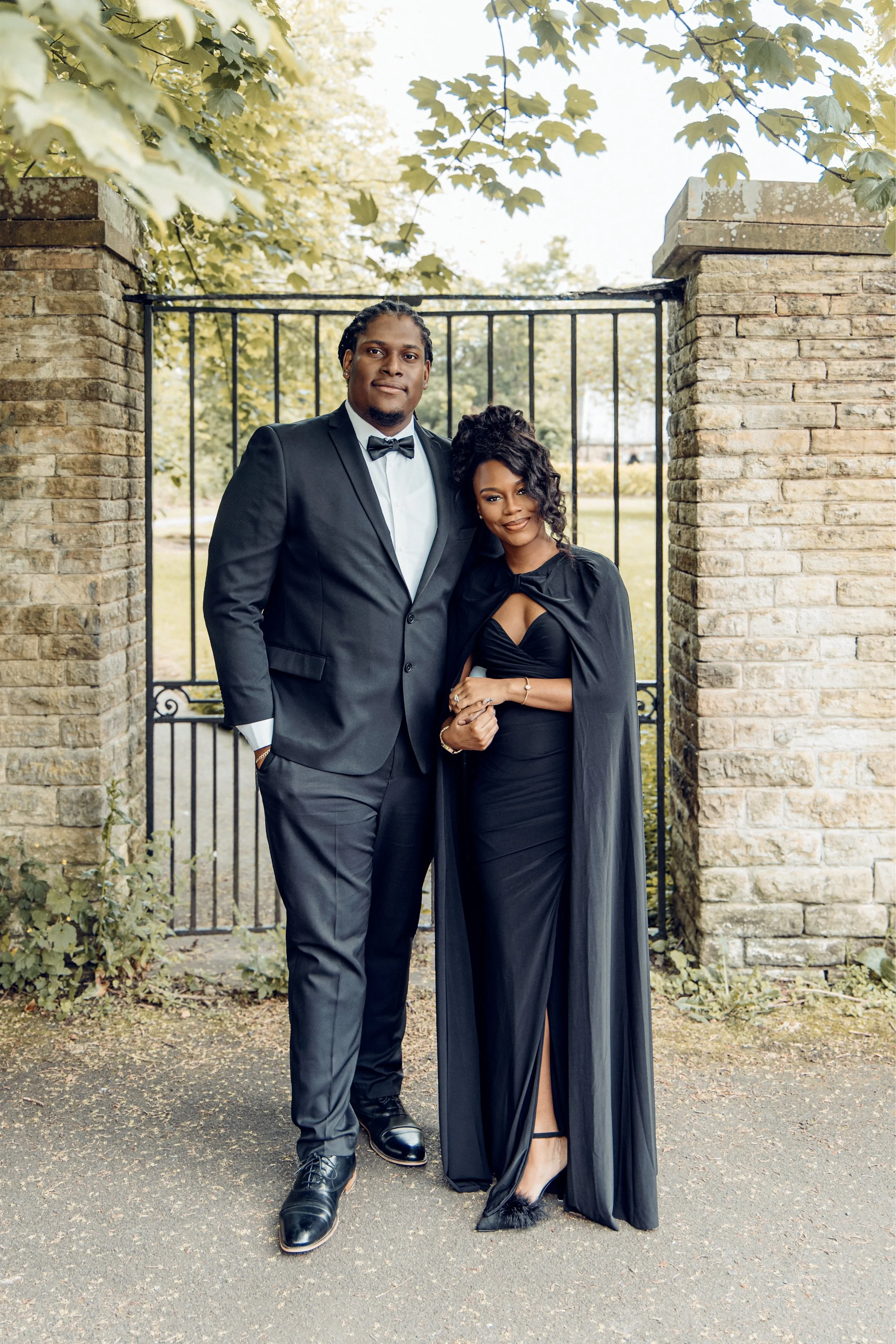 A man and woman dressed in formal attire standing outdoors in front of a brick gate with trees and foliage overhead.