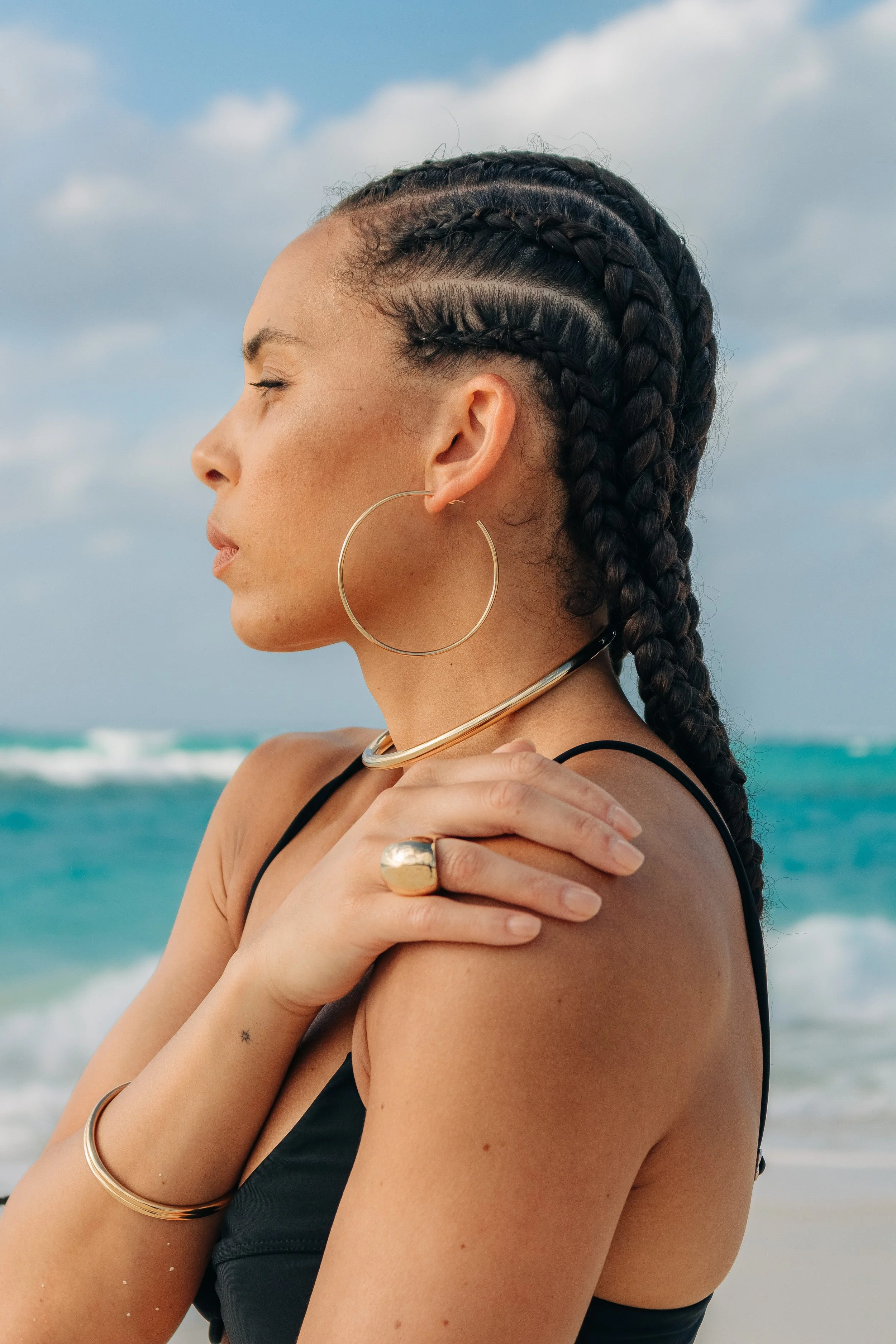 A woman with braided hair, wearing large hoop earrings, a bold ring, and gold jewelry, standing on a beach with ocean waves and cloudy sky in the background.
