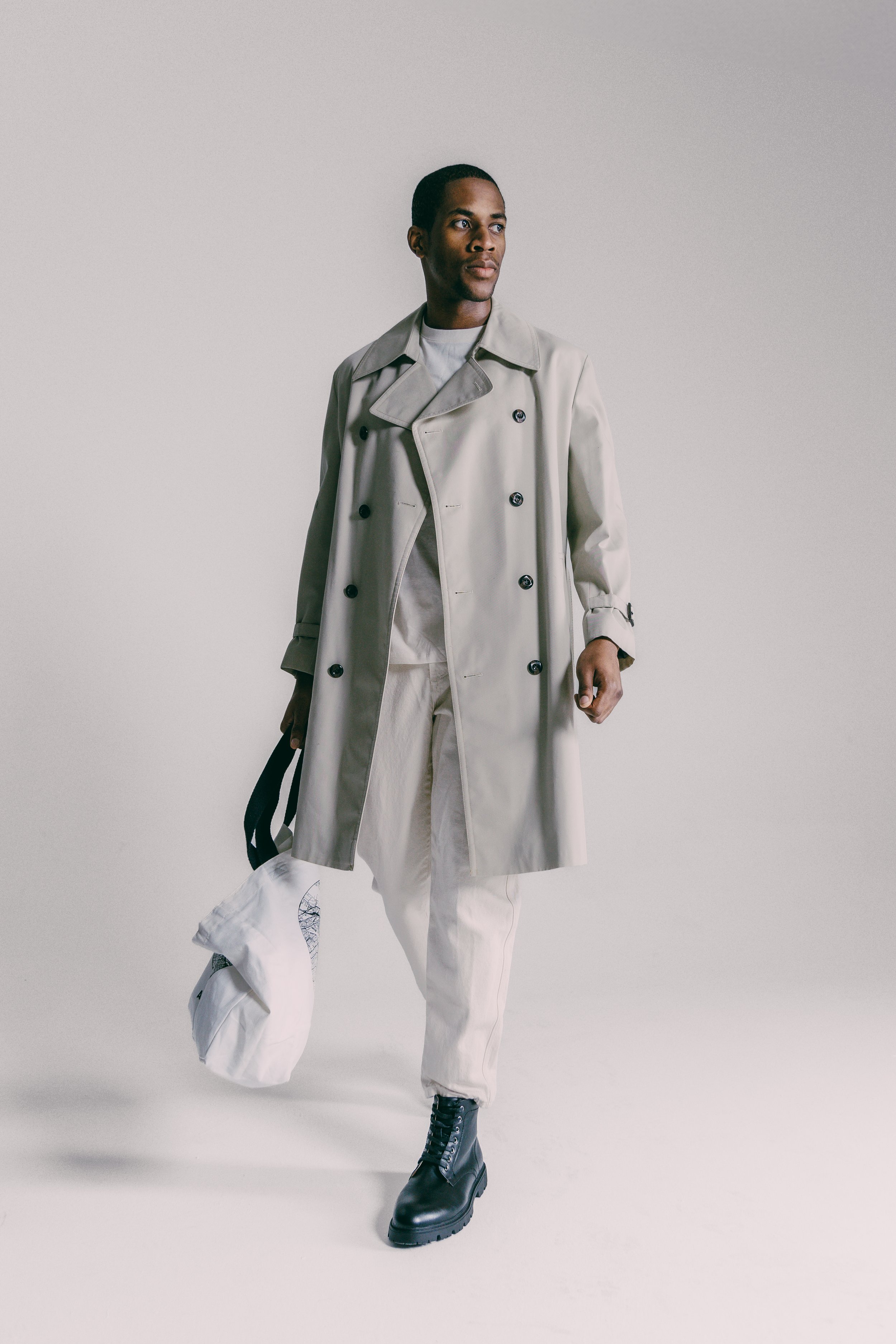 A man wearing a beige trench coat, white pants, black boots, carrying a white bag and a black strap, standing against a plain wall.