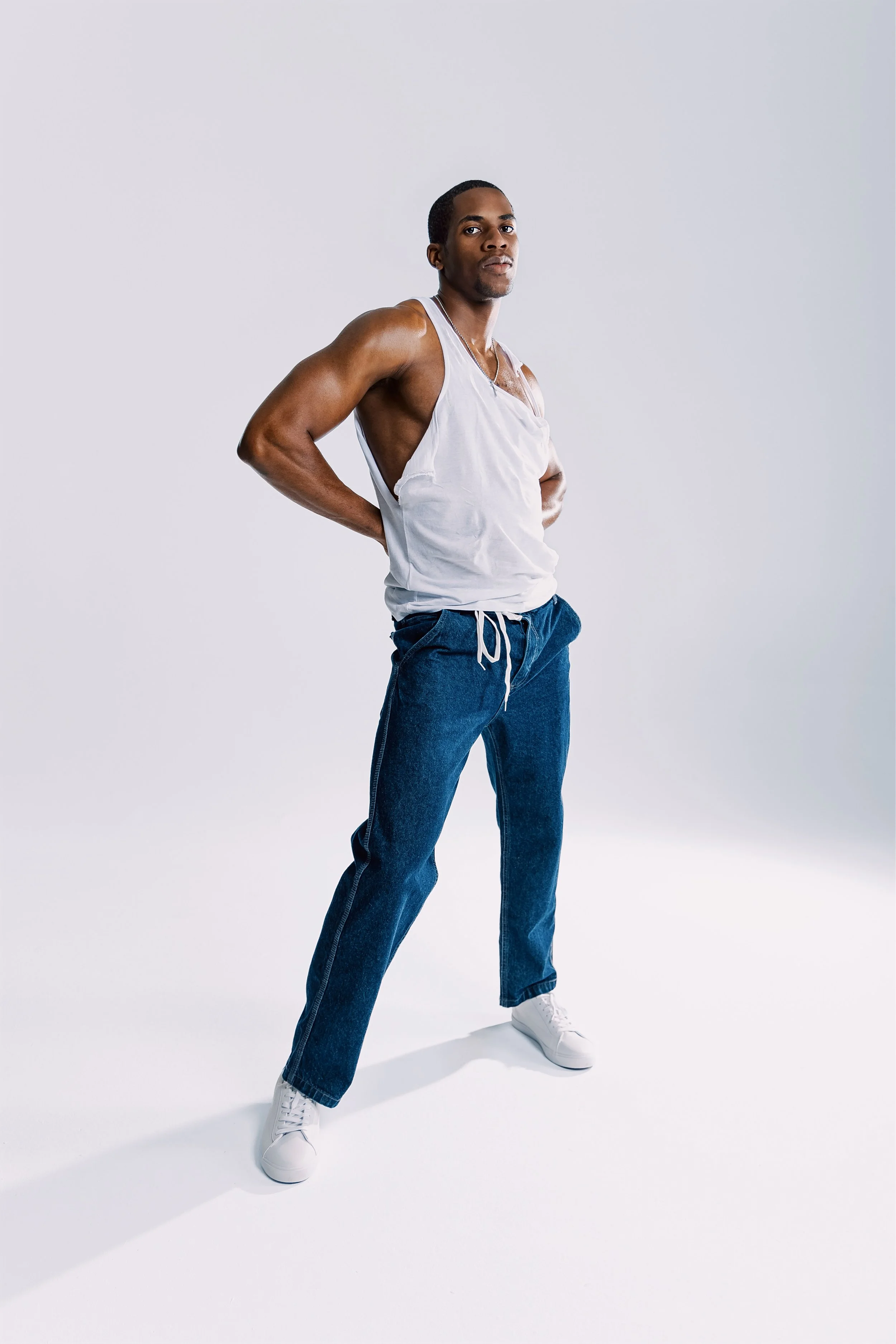 A young man standing confidently with hands behind his back, wearing a white tank top, blue jeans, and white sneakers, against a plain white background.