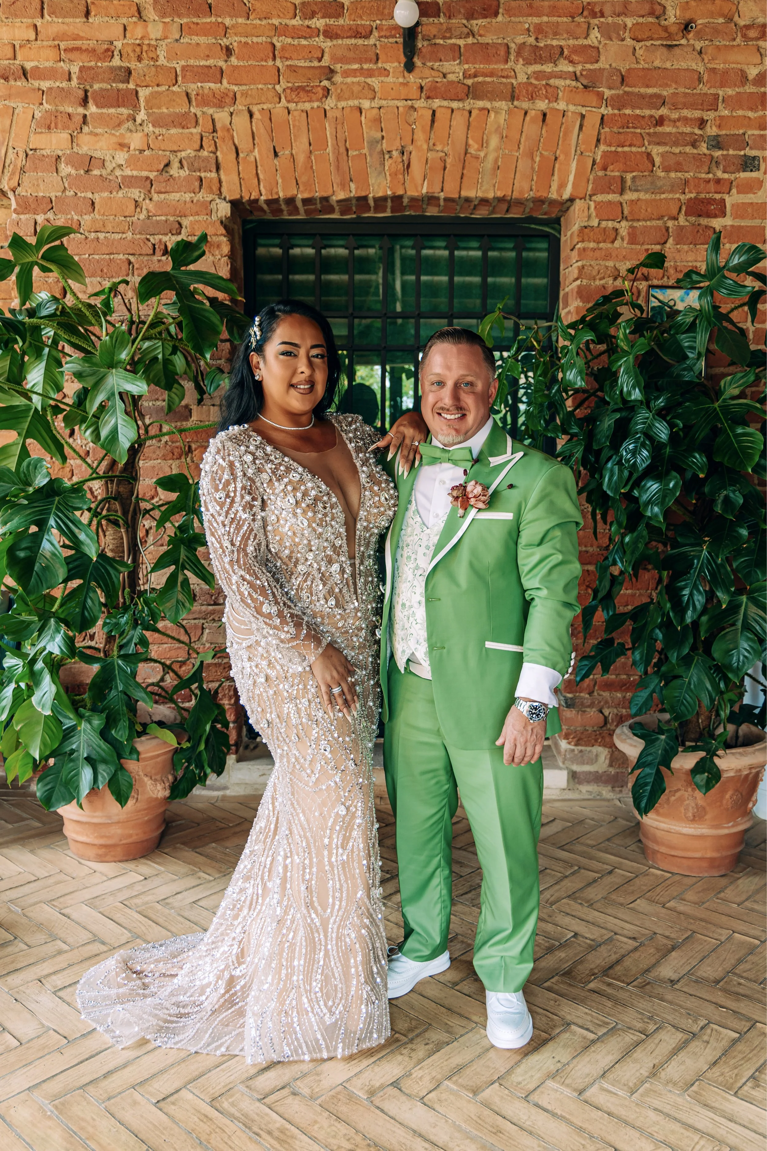 A woman in a sparkly, fitted, beige gown with a train, standing next to a man in a bright green suit, both smiling, surrounded by large green plants against a brick wall background.
