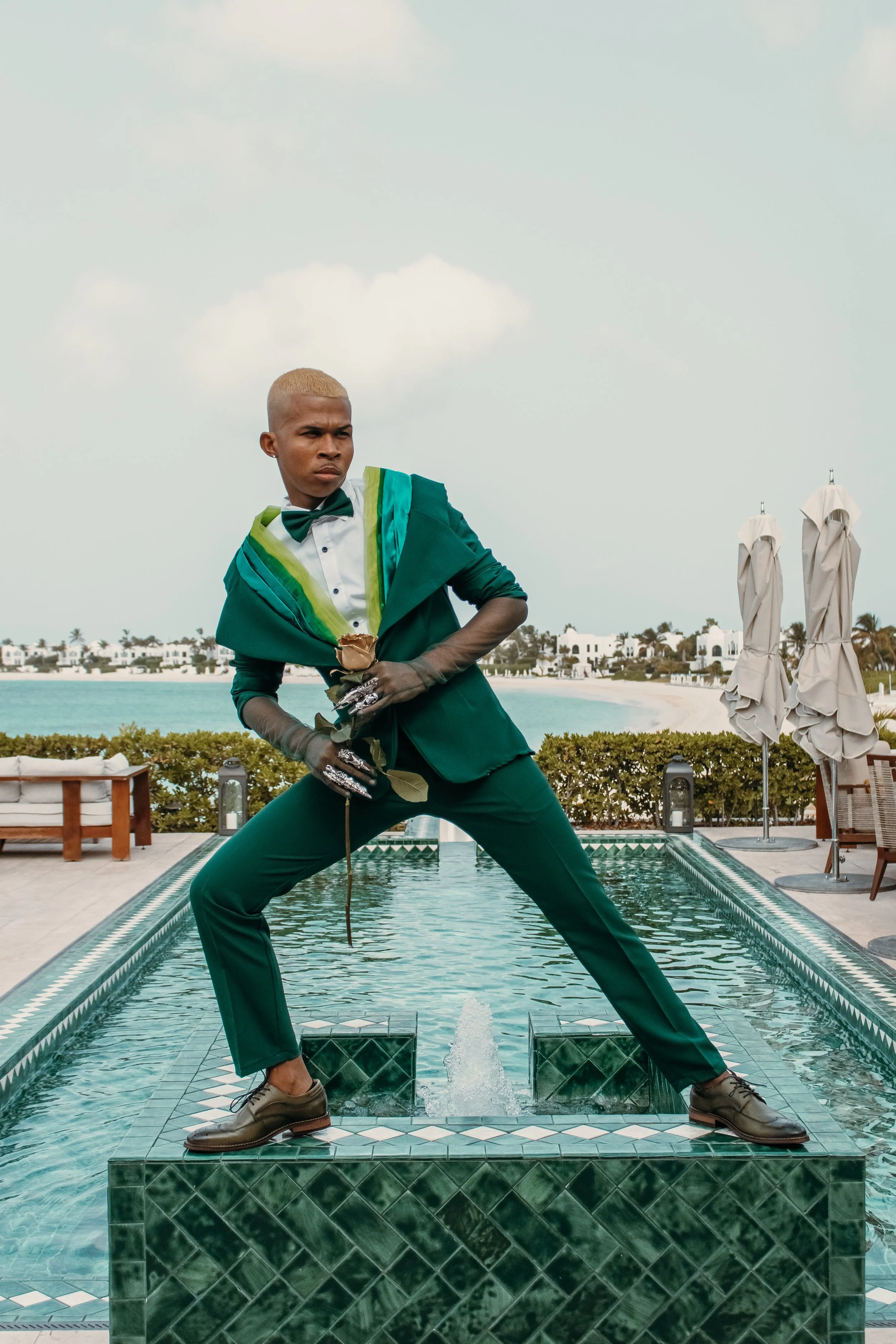 Person in formal attire standing on a fountain, holding a rose, with an ocean and beach houses in the background.