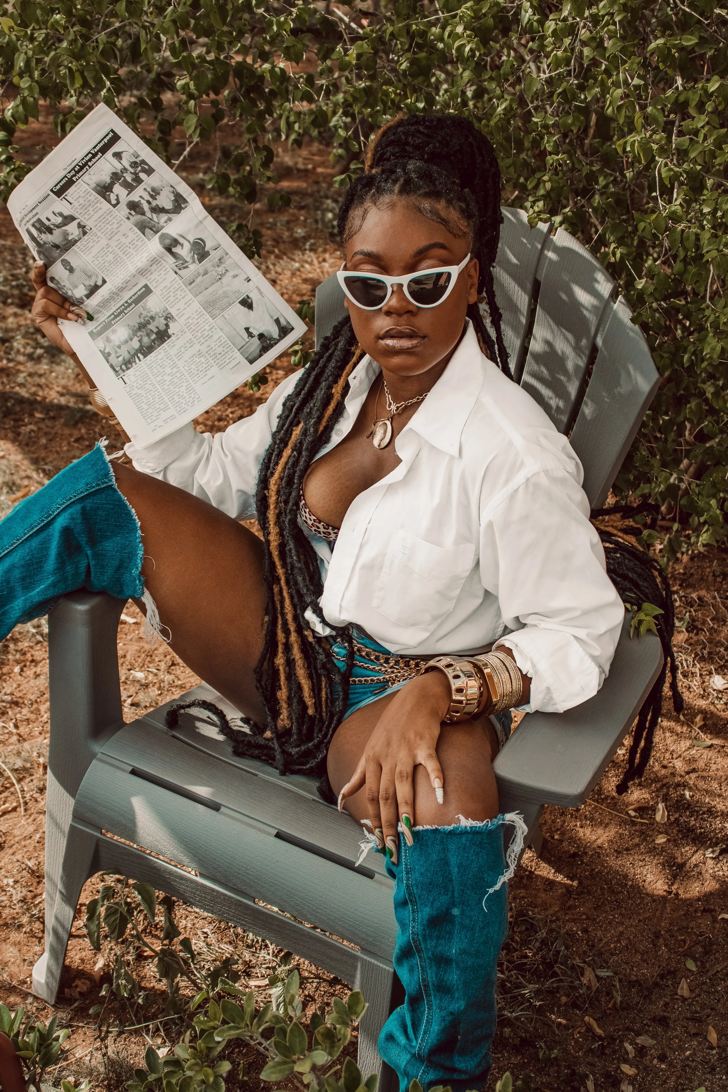 A woman with long braided hair, wearing sunglasses, a white shirt, denim shorts, and multiple bracelets, sitting on an outdoor chair and holding a newspaper.