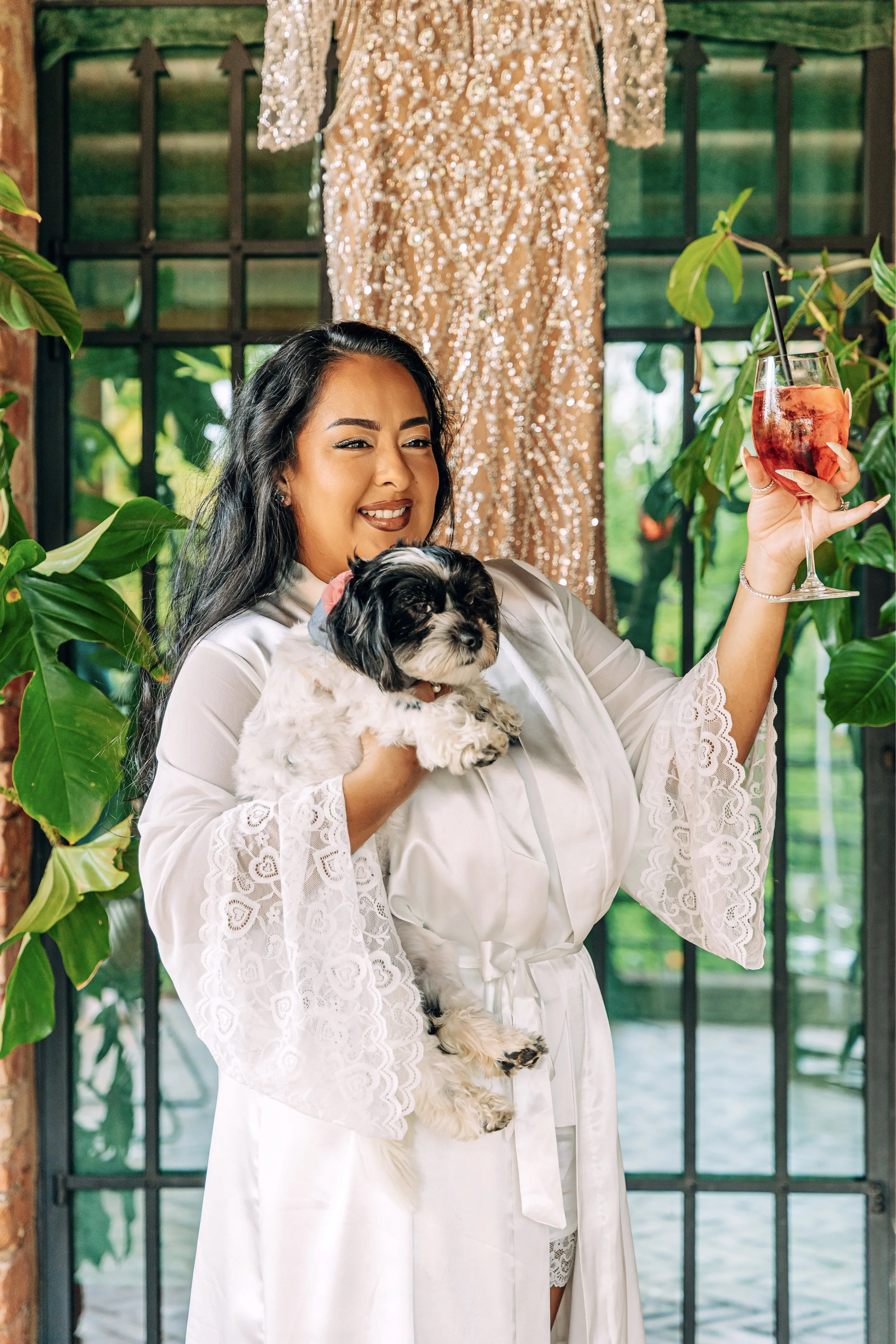 Woman in white satin robe holding a fluffy puppy and a glass of red drink, standing in front of a window with green plants and a sequined dress hanging behind her.