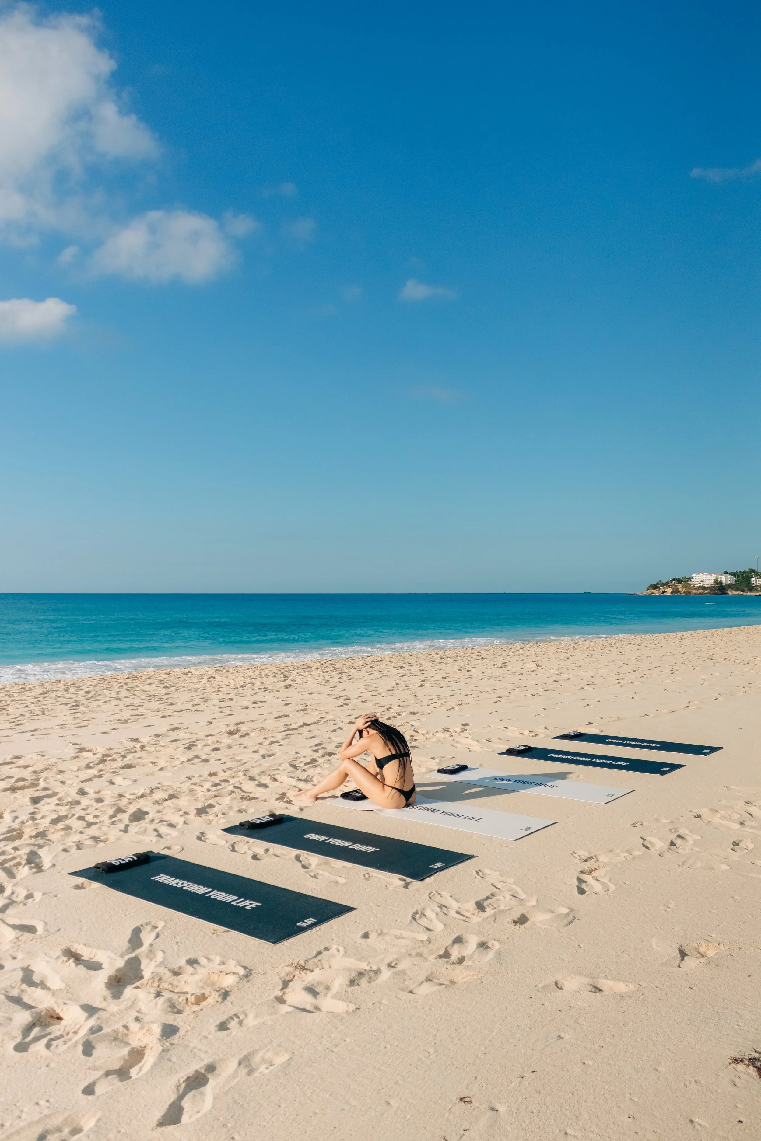 A woman in a black bikini sitting on a yoga mat on a sandy beach with the ocean and blue sky in the background.