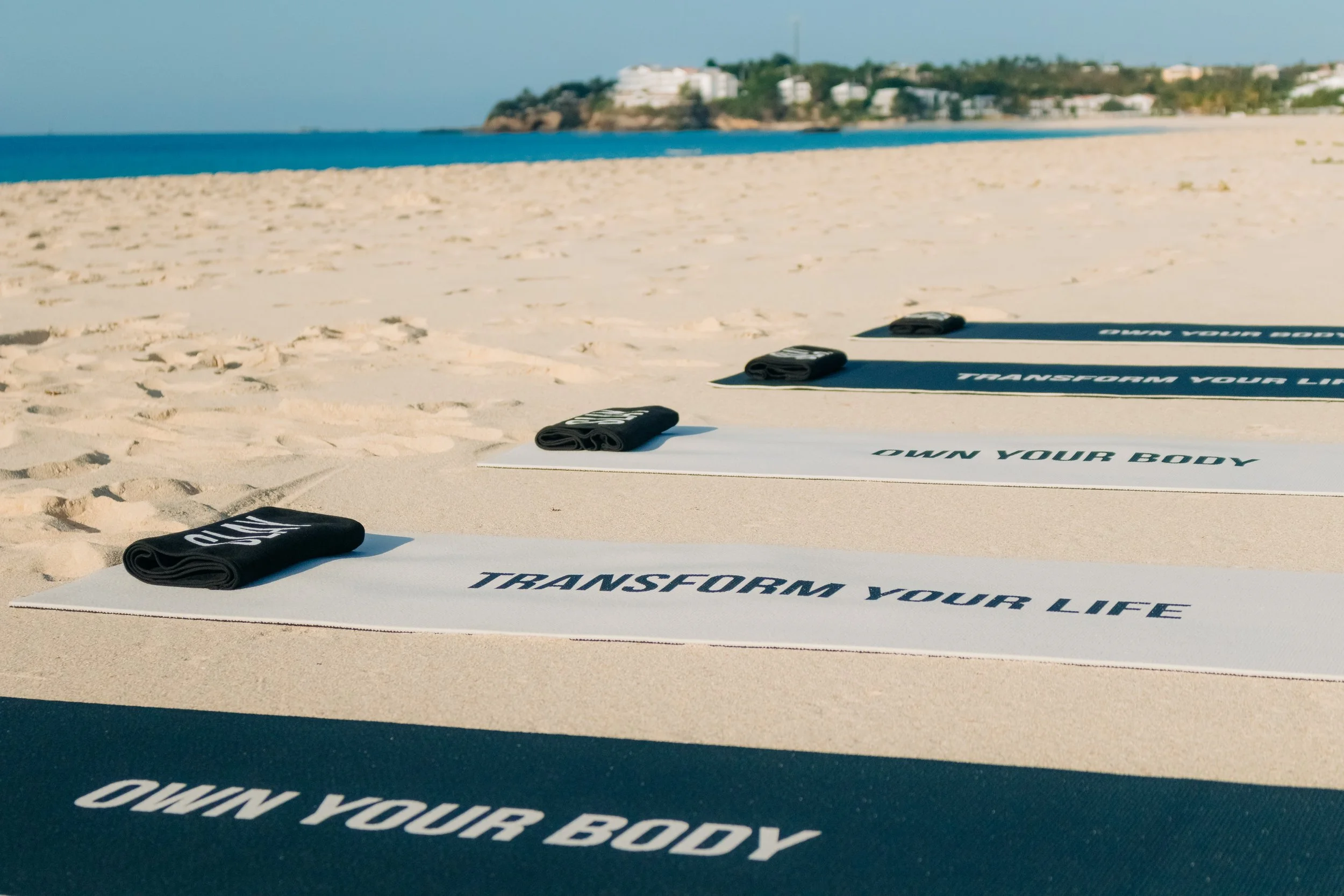 Beach yoga mats with black towels on a sandy beach facing the ocean and a distant shoreline with buildings.