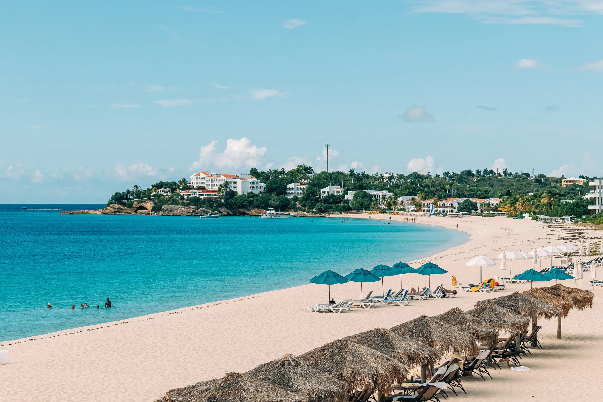 A tropical beach with white sand, blue waters, and thatched umbrellas. There are people swimming and lounging in the water, and buildings with red roofs and lush greenery on a hillside in the background, under a blue sky with a few clouds.
