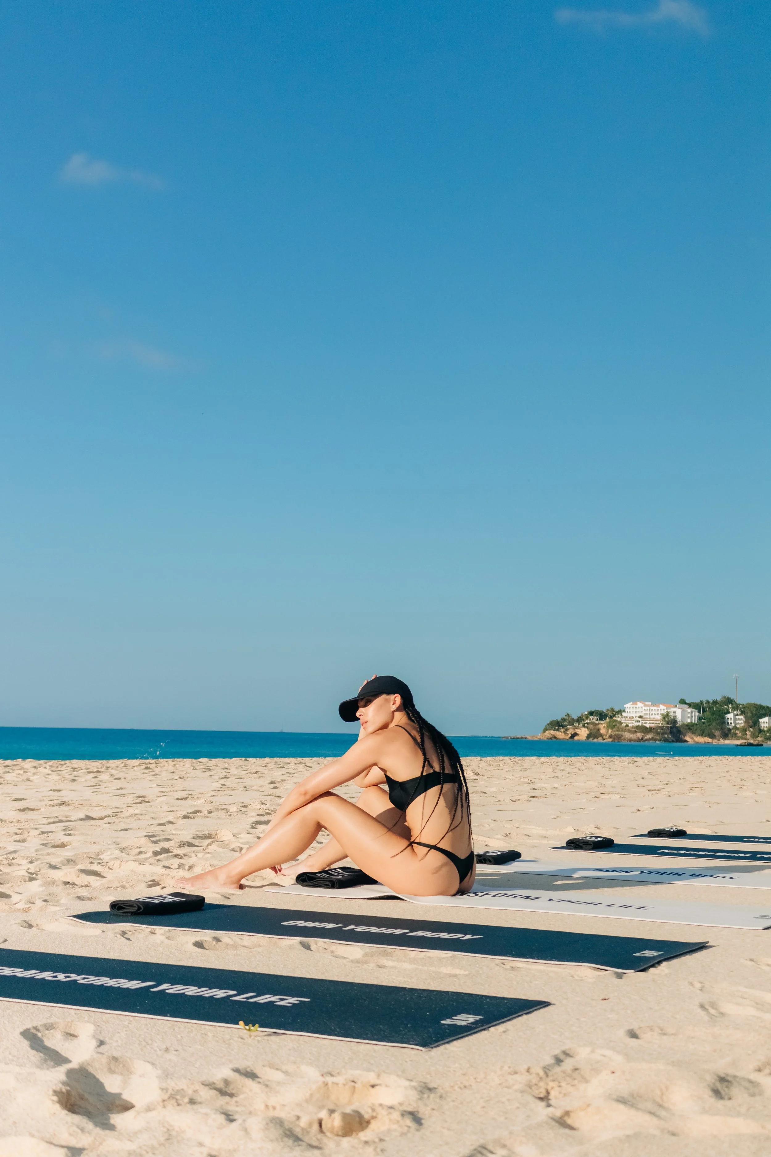 A woman in a black swimsuit and cap sitting on a beach towel at the sandy beach with the ocean and a distant coastline in the background.