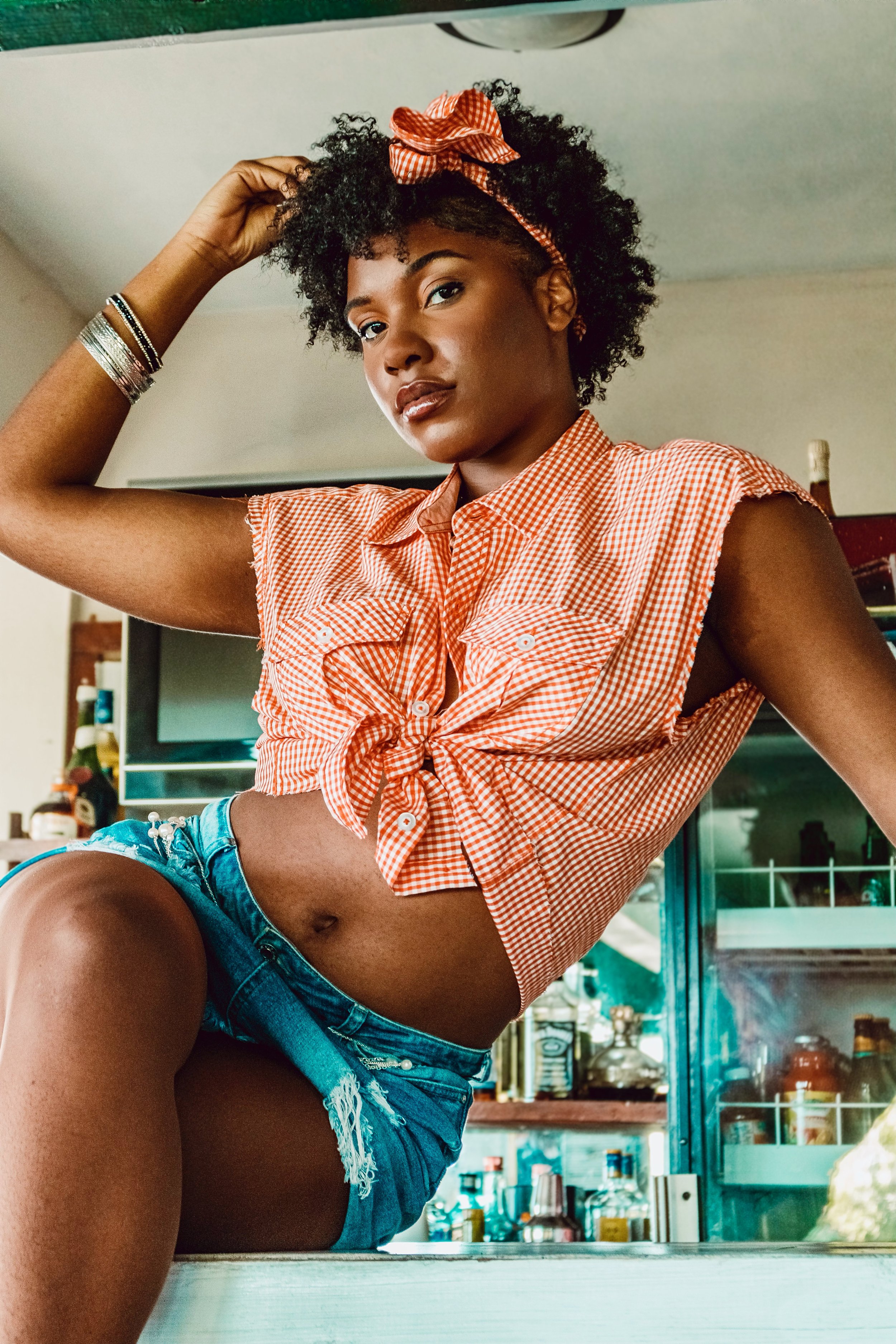 A young woman with curly black hair wearing a red and white checkered sleeveless shirt tied at the waist, denim shorts, and a matching headband with a bow posing indoors. She has several bracelets on her right wrist and is touching her hair with her 