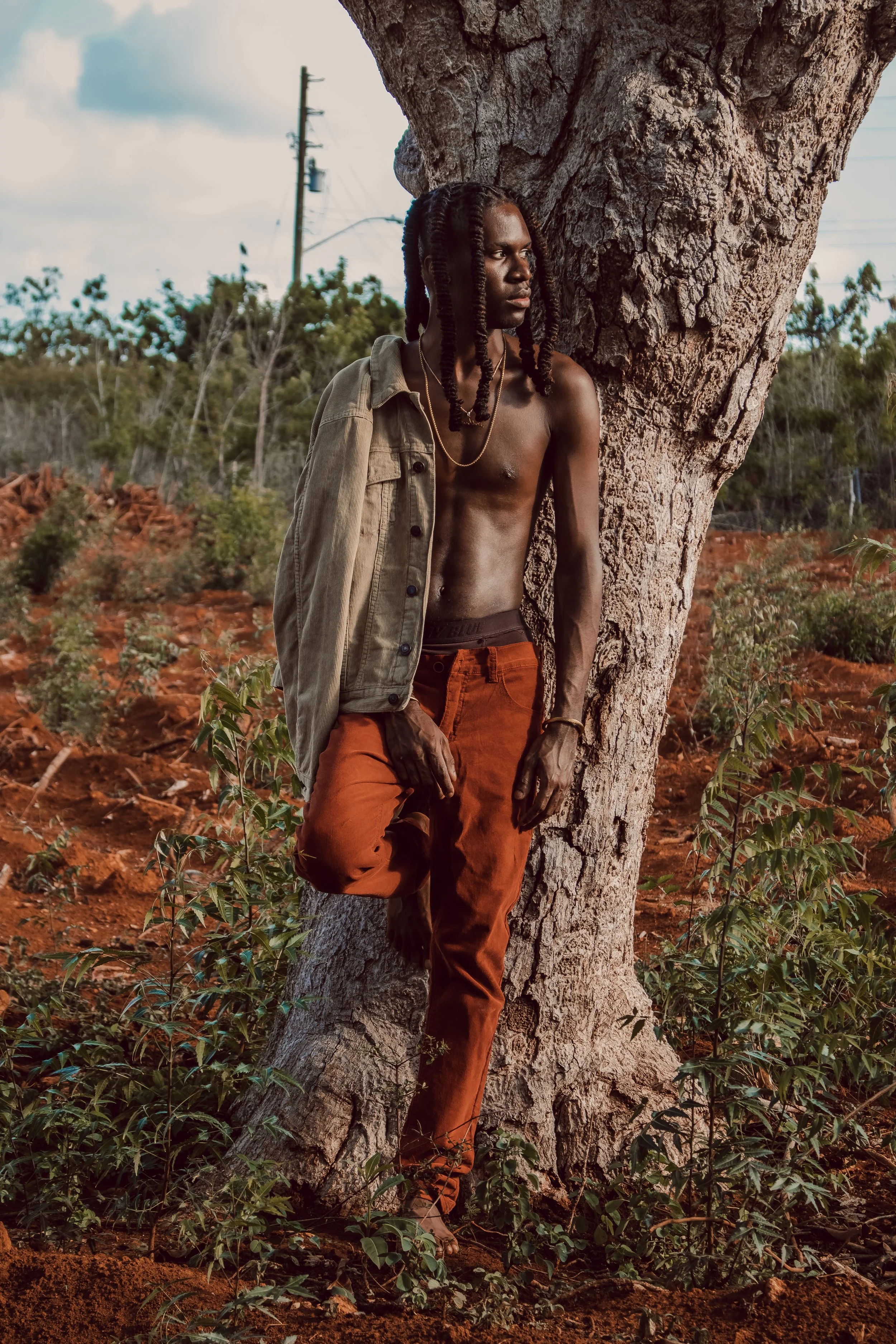 A shirtless man with dreadlocks leaning against a tree in a natural landscape, wearing orange pants and a beige jacket draped over his shoulders, looking to the right.