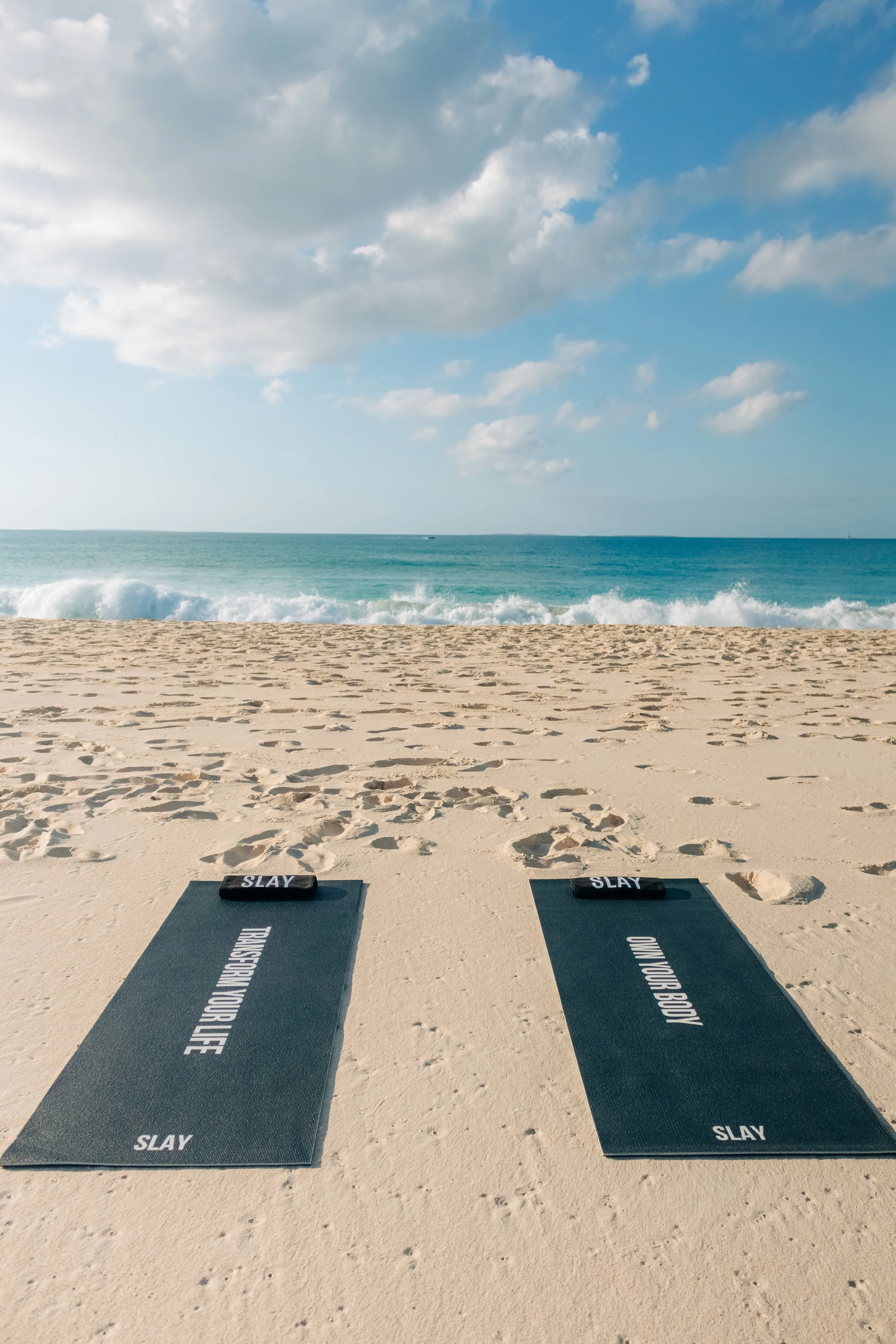Two yoga mats on a sandy beach facing the ocean with waves and a cloudy sky.