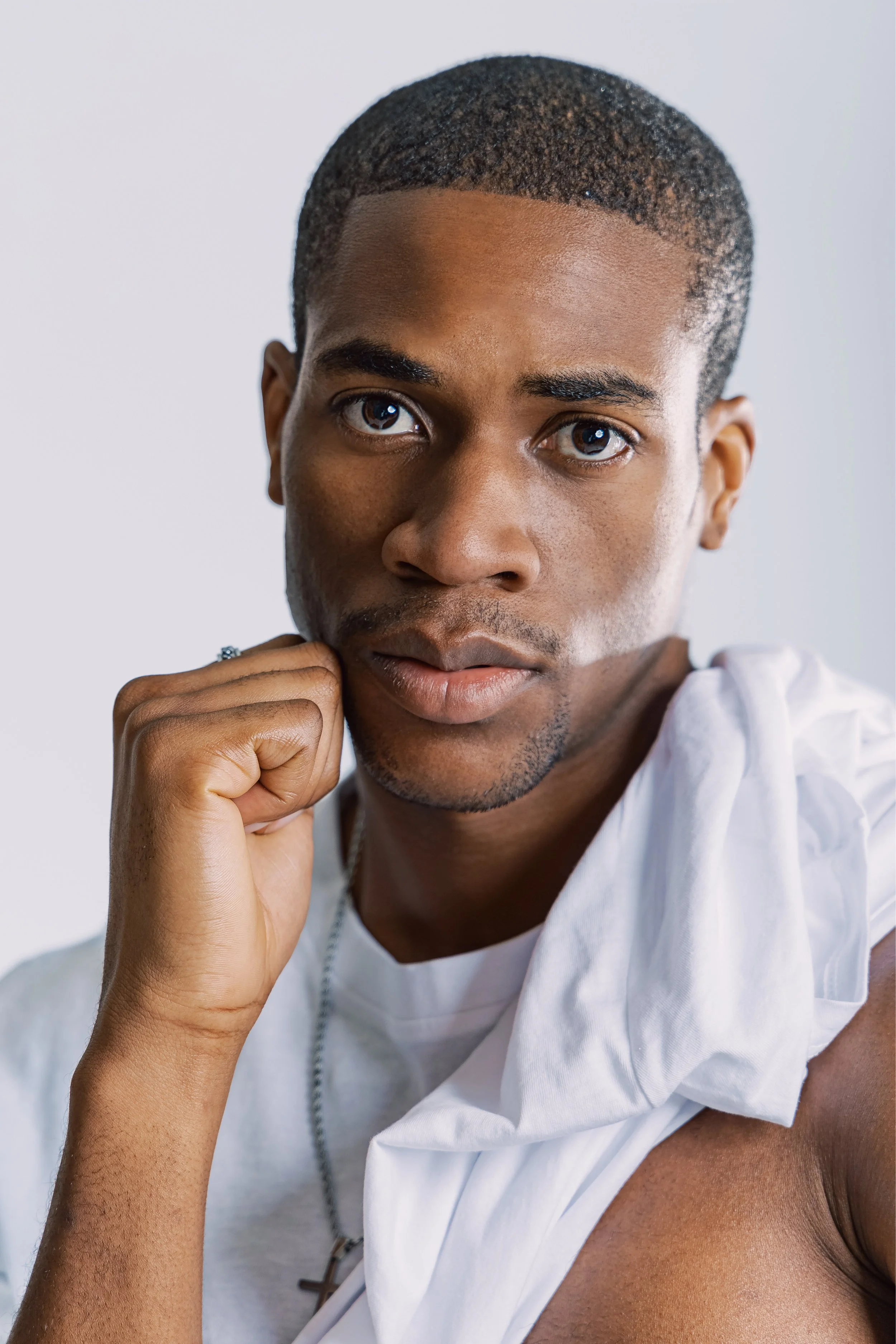 Close-up portrait of a young man with short hair and a serious expression, resting his chin on his hand, wearing a white garment draped over his shoulder.